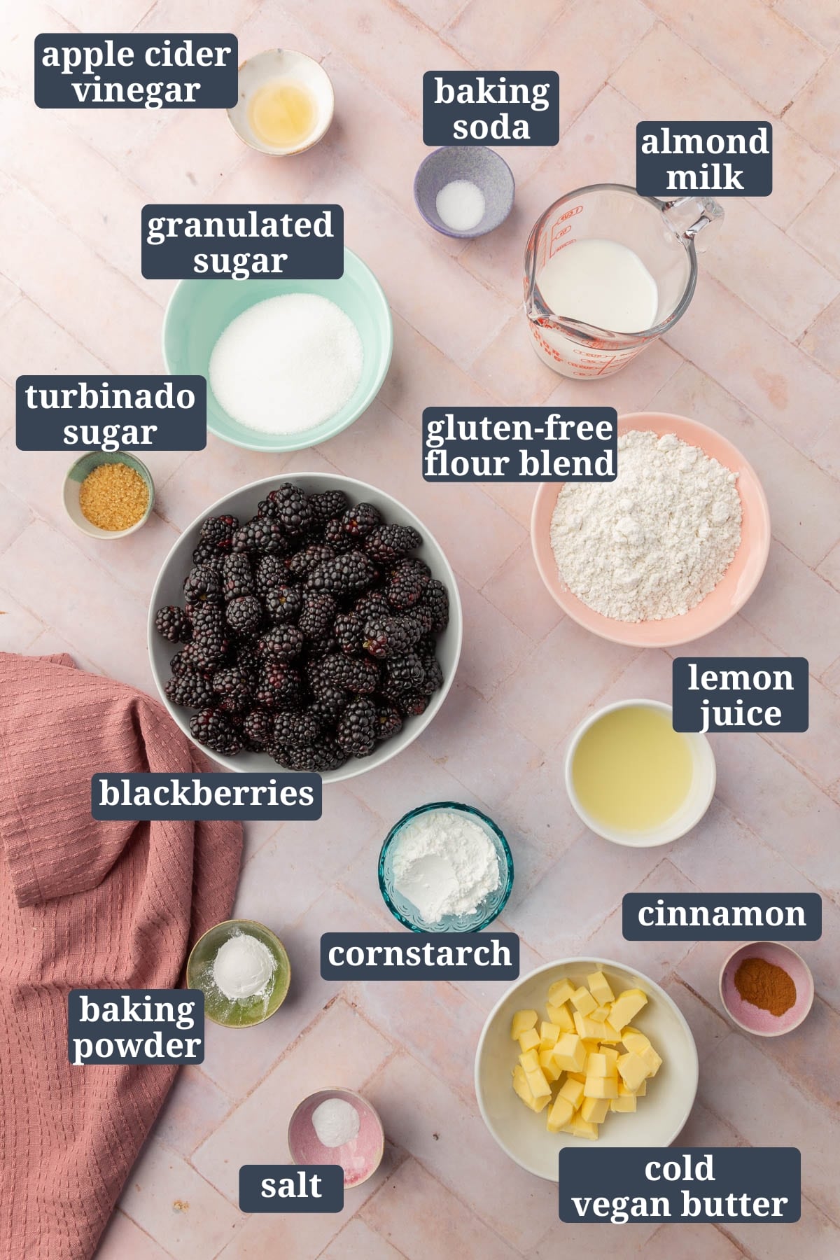 An overhead view of ingredients in small bowls to make gluten-free blackberry cobbler, including apple cider vinegar, salt, almond milk, granulated sugar, turbinado sugar, blackberries, gluten-free flour, lemon juice, cornstarch, baking powder, baking soda, vegan butter, and cinnamon with text overlays over each ingredient.