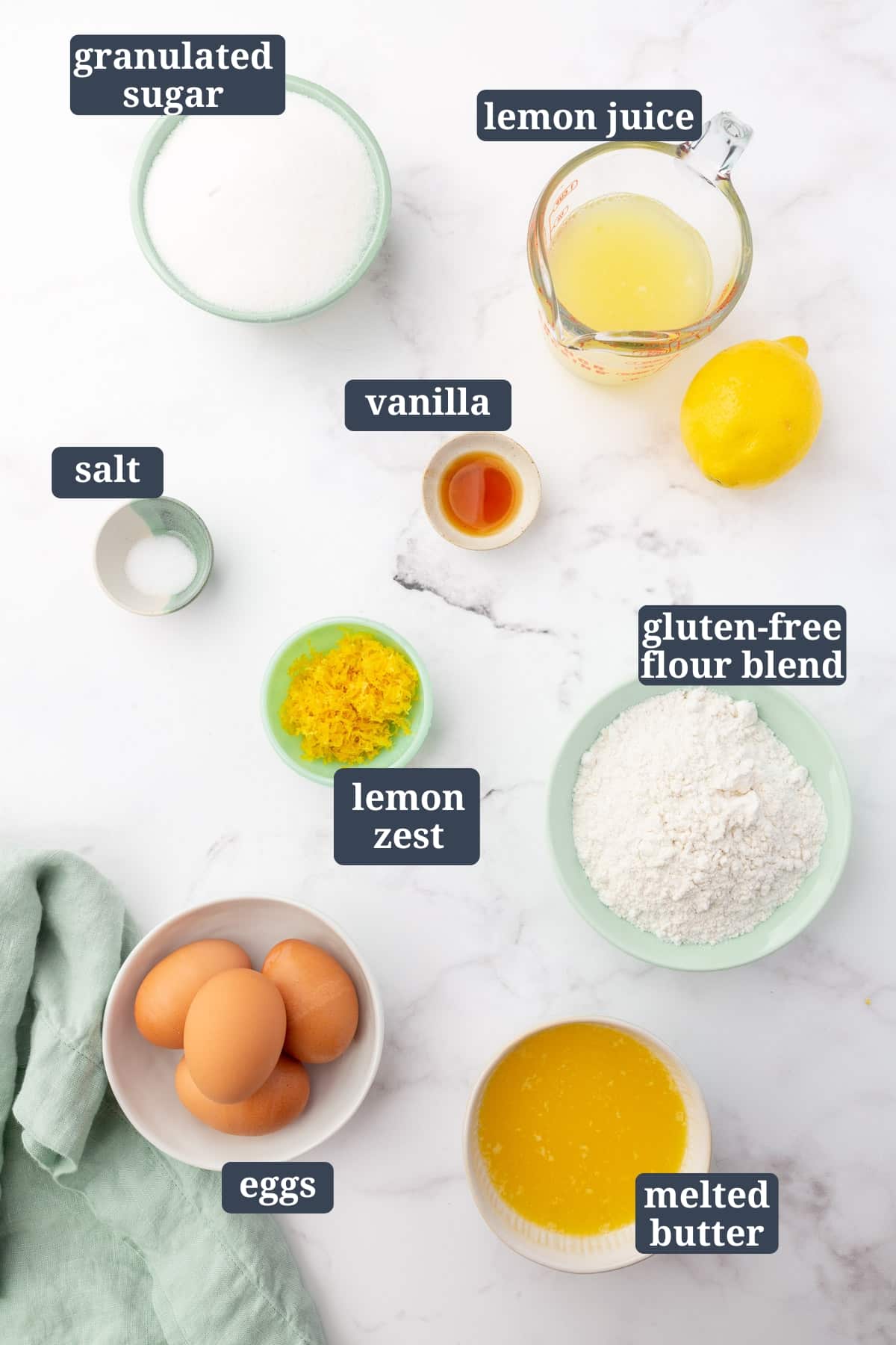 An overhead view of ingredients to make gluten free lemon bars, including granulated sugar, lemon juice, vanilla, salt, lemon zest, gluten-free flour, eggs, and melted butter with text overlays over each ingredient.