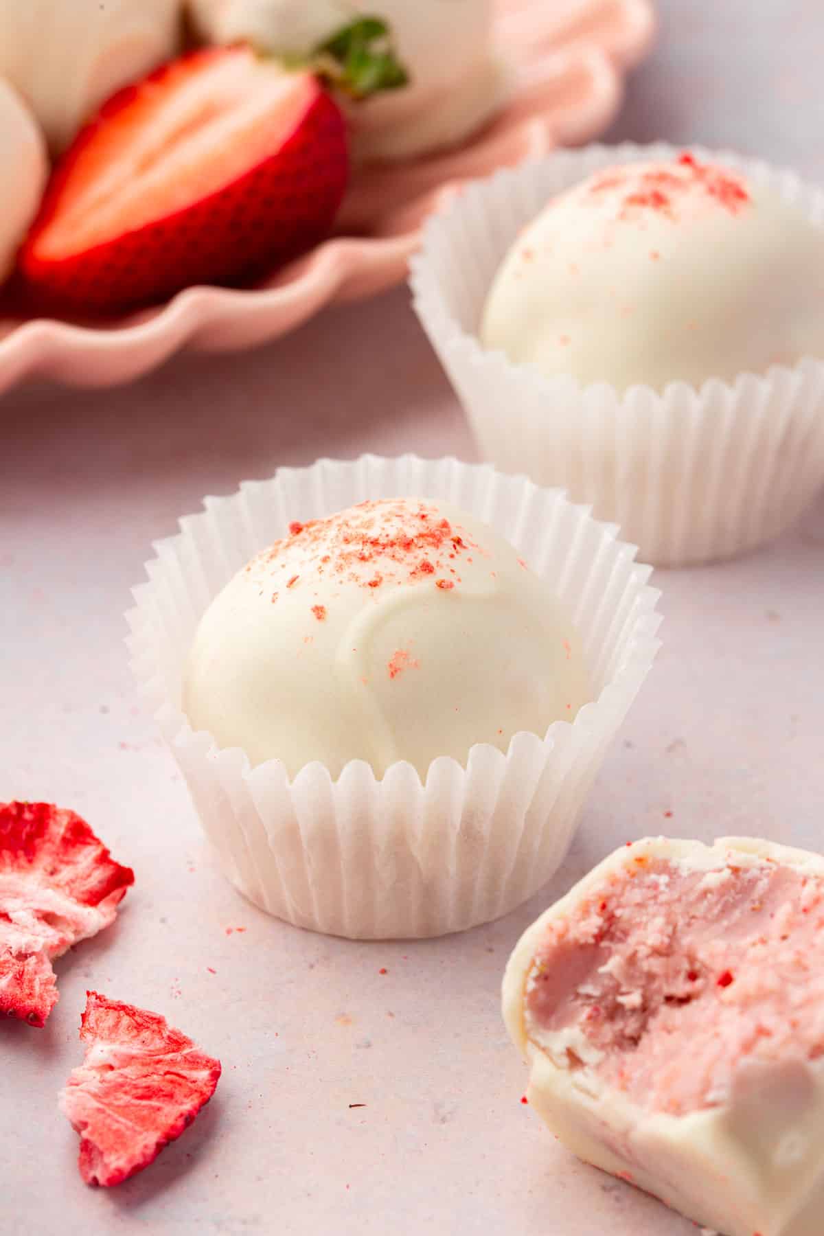 Two strawberry white chocolate truffles in paper cups with freeze-dried strawberries, fresh strawberries and additional truffles in the background.
