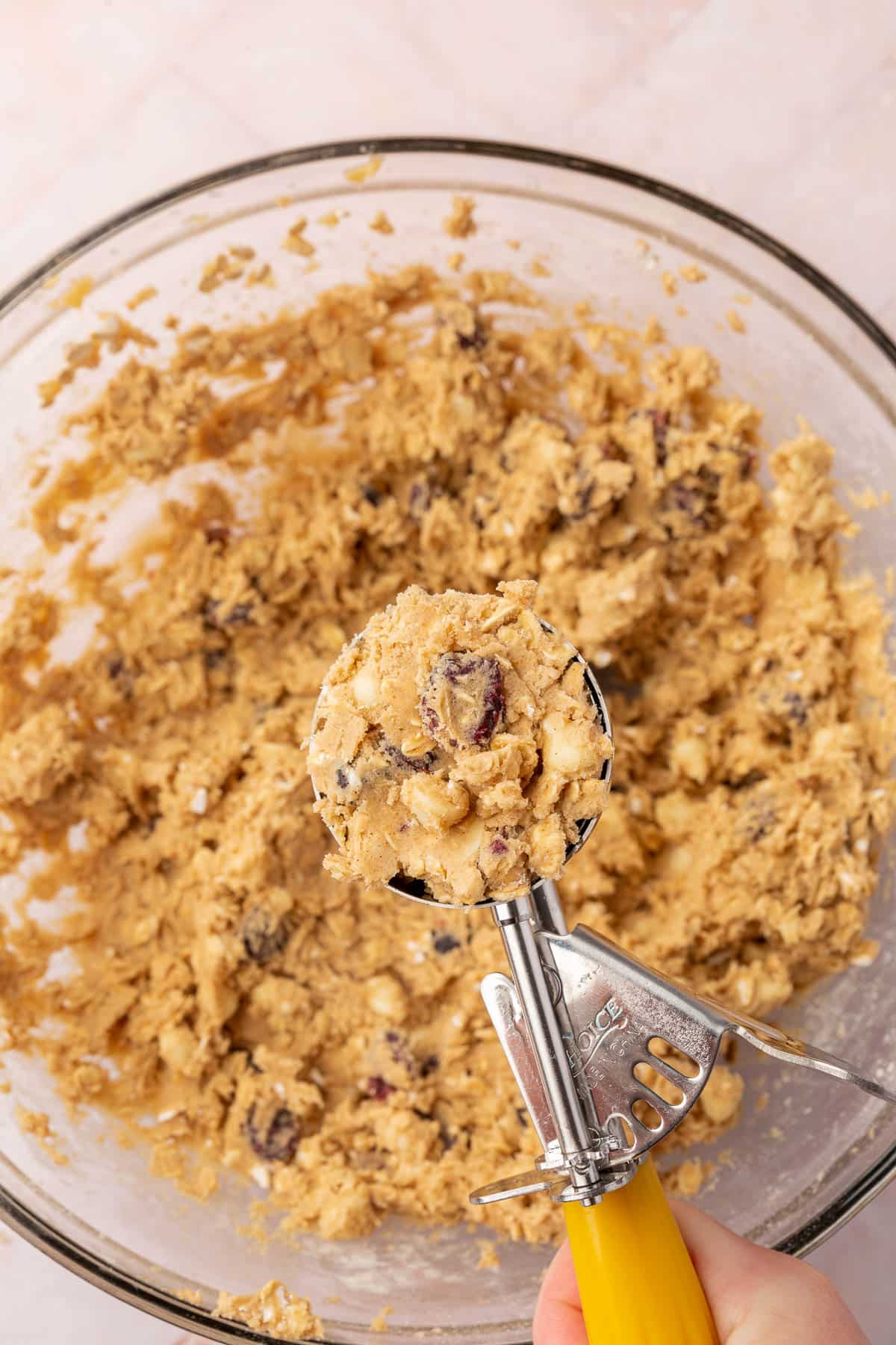 A yellow portion scoop of oatmeal cranberry white chocolate cookie dough over a large bowl of dough.