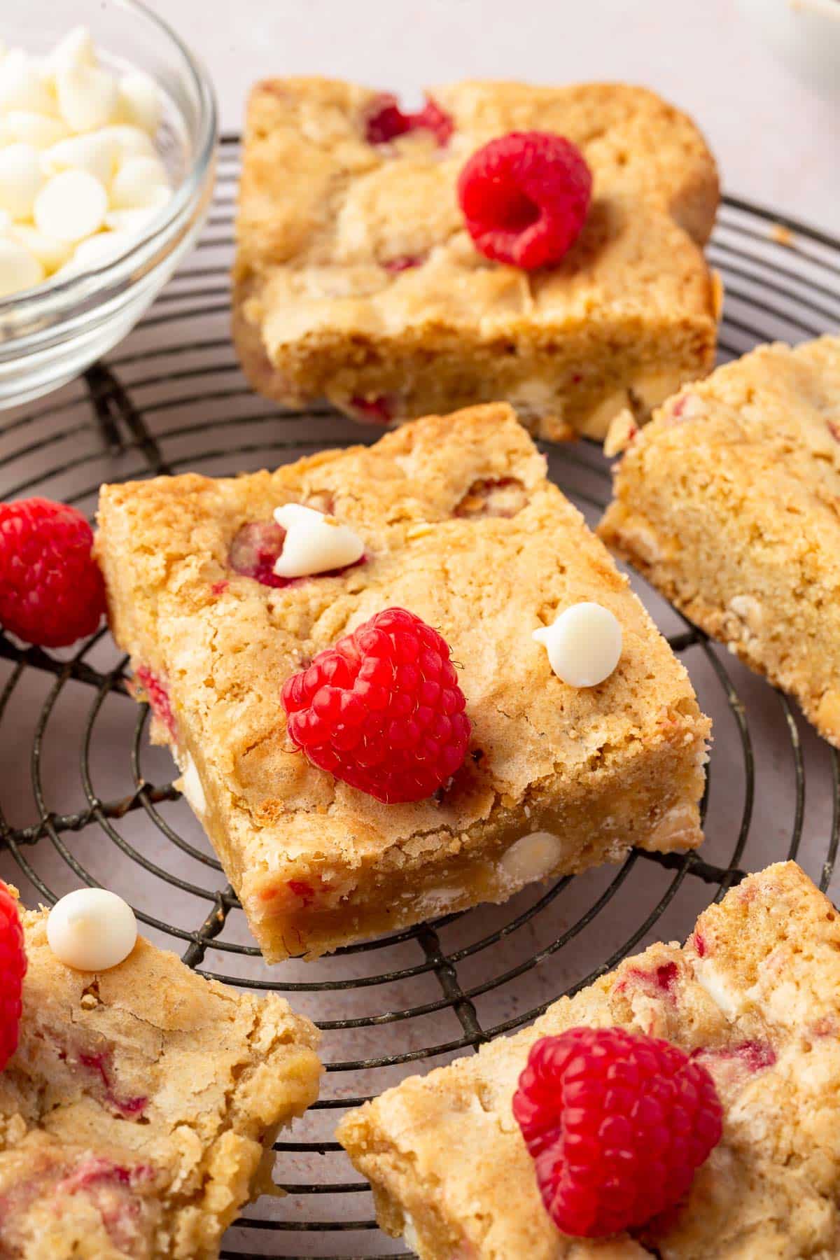 Multiple gluten-free white chocolate and raspberry blondies on a cooling rack with a bowl of white chocolate chips and fresh raspberries decorated on top of the blondies.