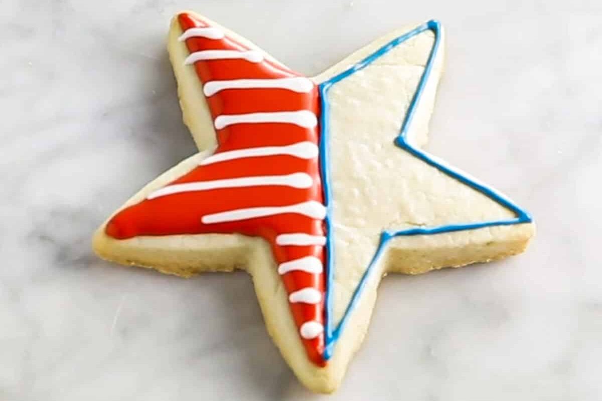 A star shaped sugar cookie that is being decorated with red royal icing with white stripes and an outline of blue royal icing on the other half of the cookie.