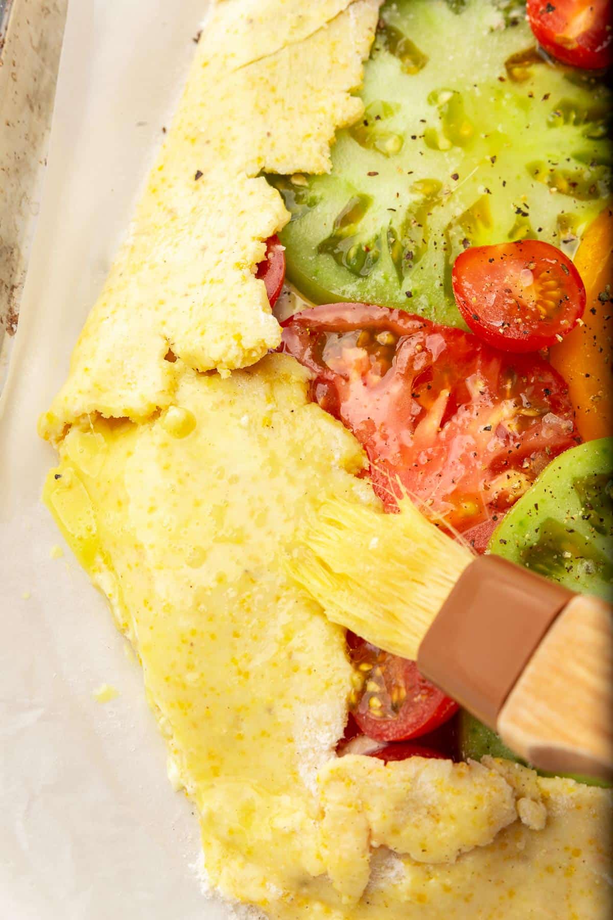 A pastry brush spreading egg wash on the gluten-free galette dough edges of a tomato tart.