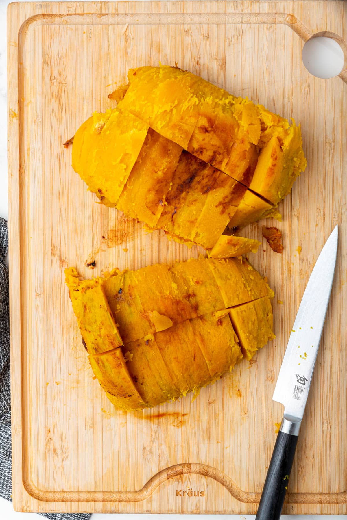 A cutting board with two roasted pumpkin halves that have been cut into chunks with a knife next to it.
