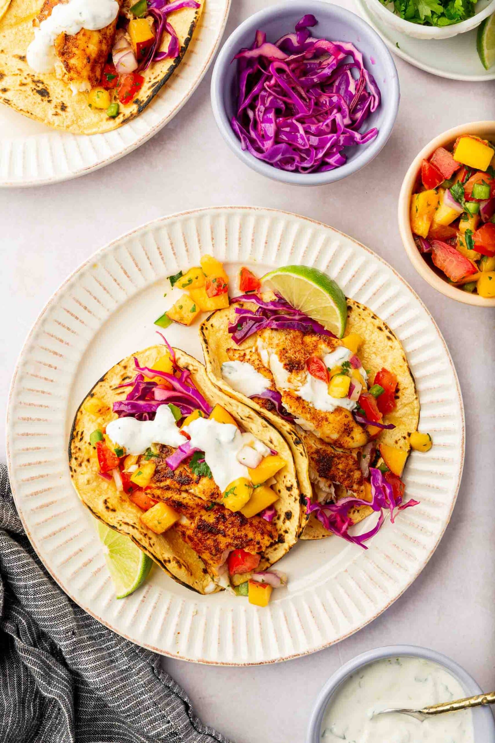 Two fish tacos on charred corn tortillas topped with sour cream, fruit salsa, and red cabbage with bowls of slaw, fruit salsa, and crema to the side.