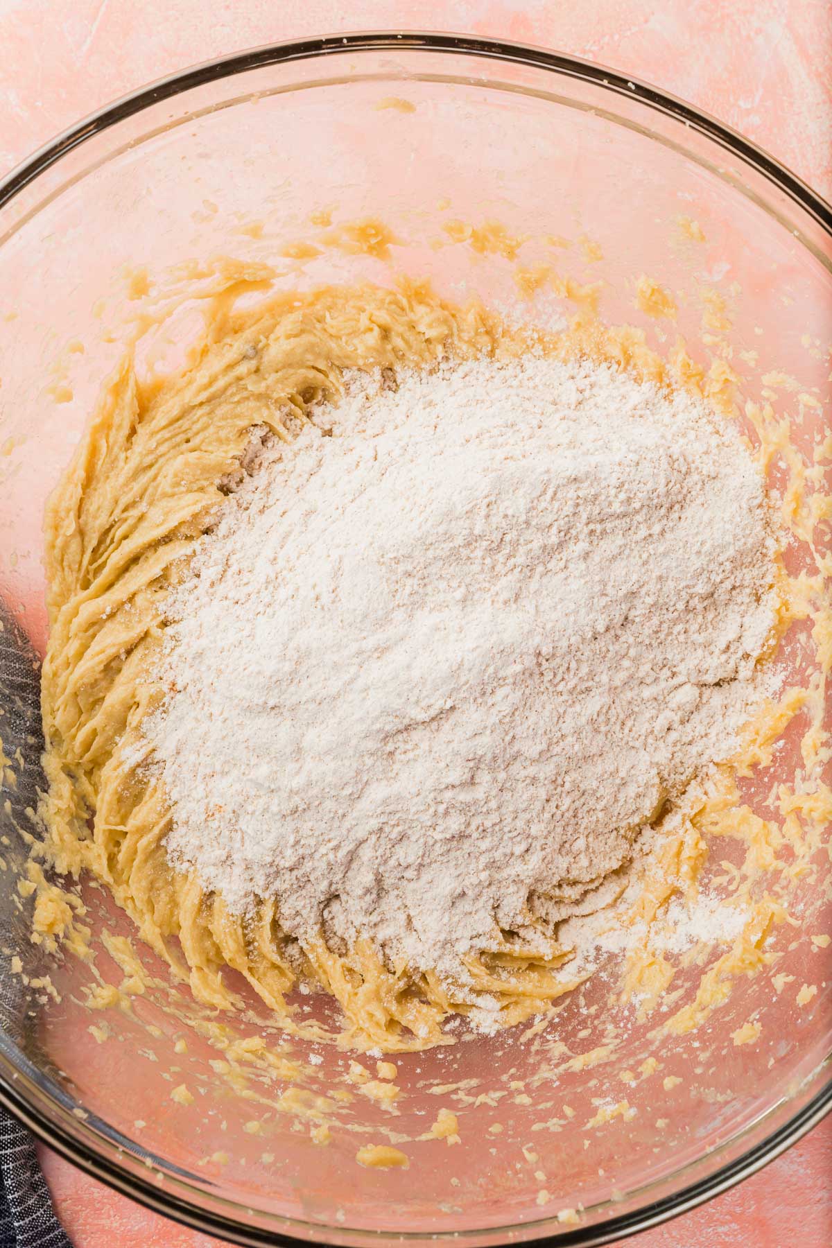 A glass mixing bowl of gluten-free cookie dough topped with gluten free flour on a pink table.