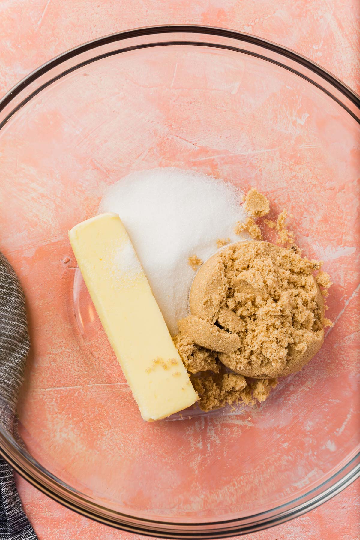 A glass mixing bowl with a stick of butter, granulated sugar and brown sugar in it before mixing together.