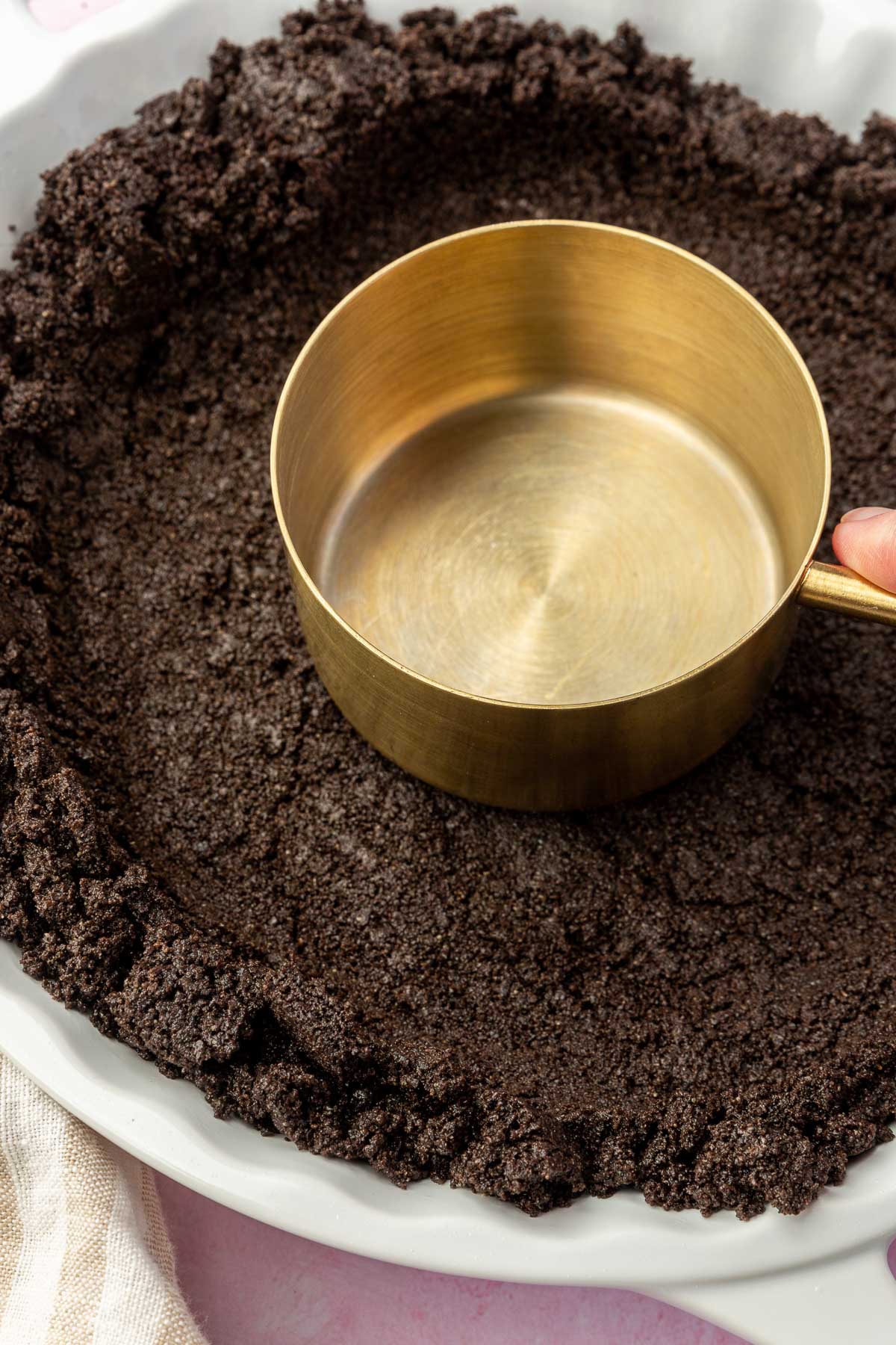 A hand pressing a gold measuring cup into a gluten-free Oreo pie crust to form an even layer in a white tart pan.