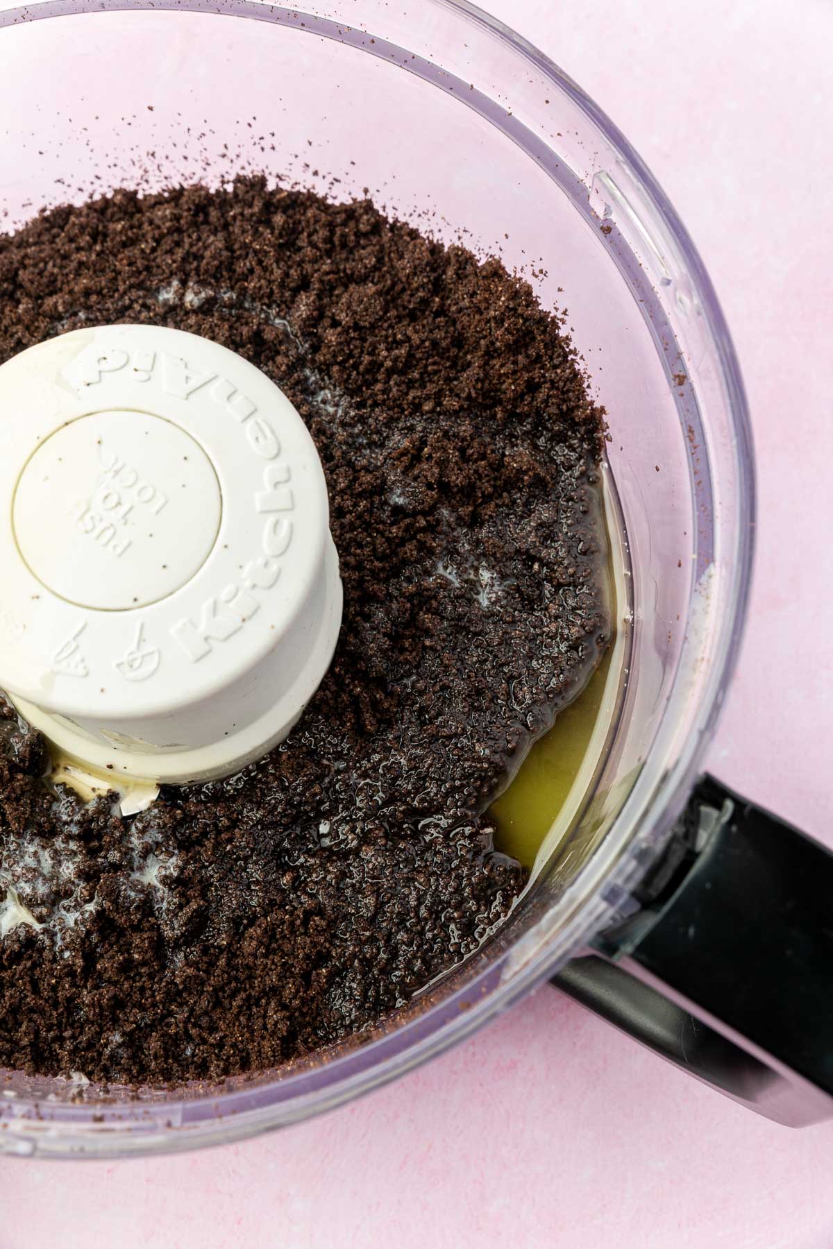 A food processor with gluten-free Oreo crumbs topped with melted butter before mixing together.