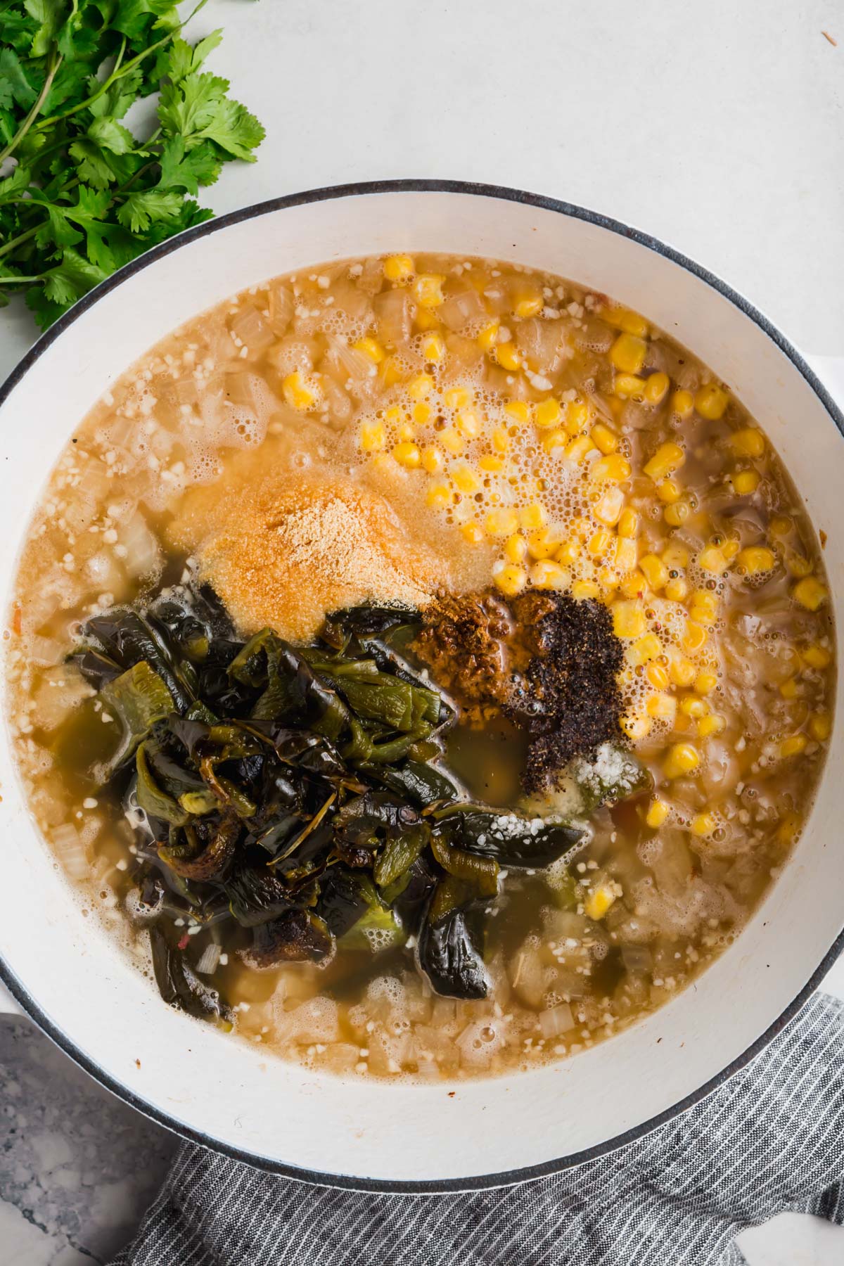 An overhead view of a white dutch oven with corn, onions, spices, roasted poblano peppers and chicken broth.