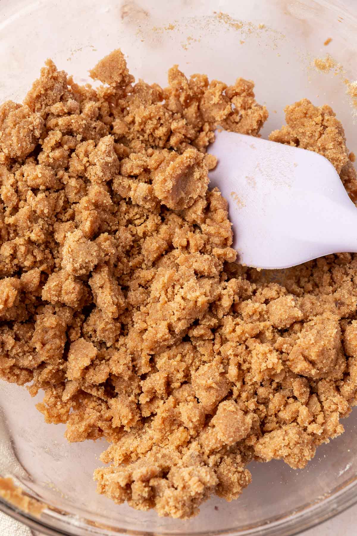 A glass mixing bowl with a gluten-free streusel topping that has been mixed together with a purple rubber spatula.