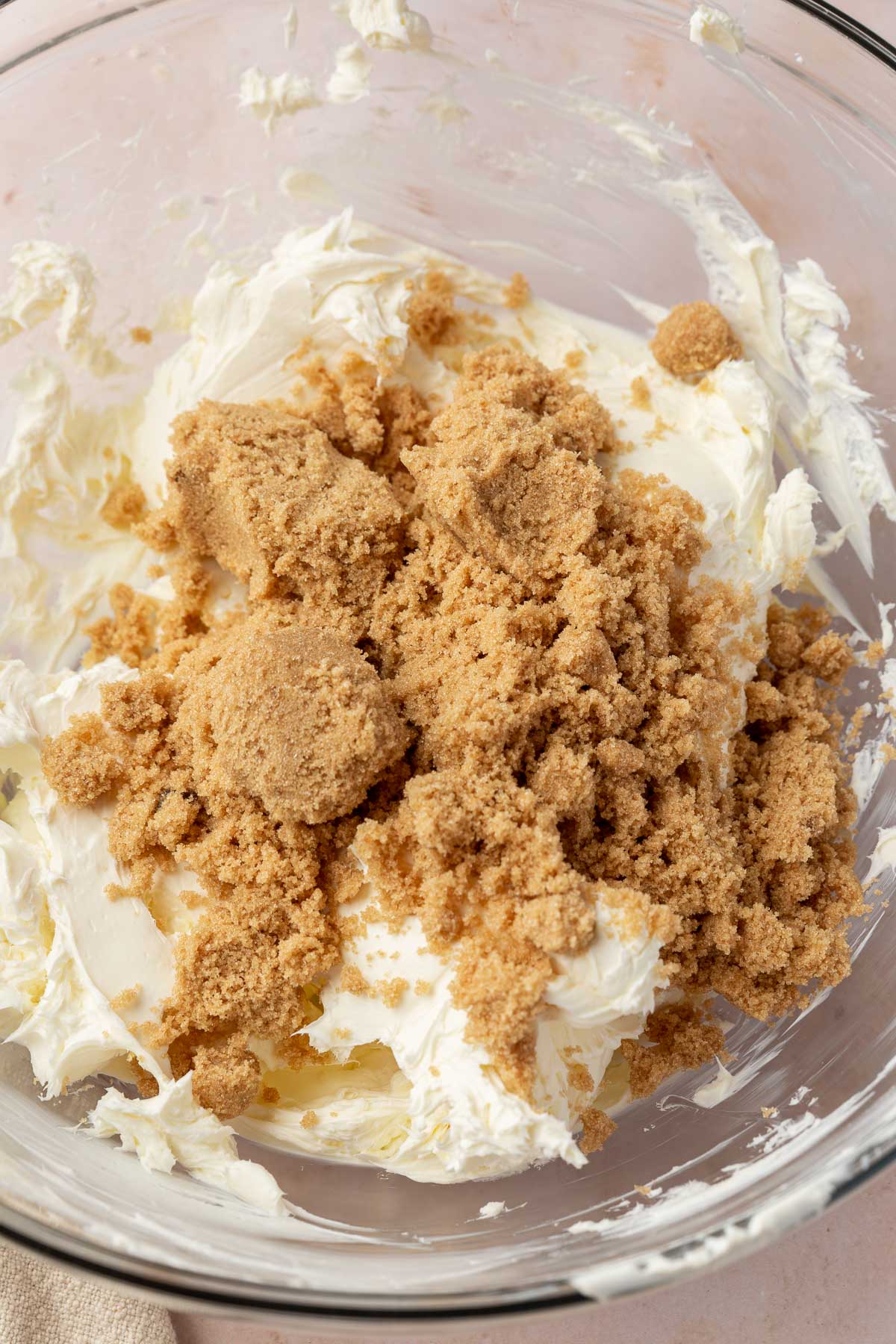 A glass mixing bowl with softened cream cheese that has been topped with brown sugar before mixing together.