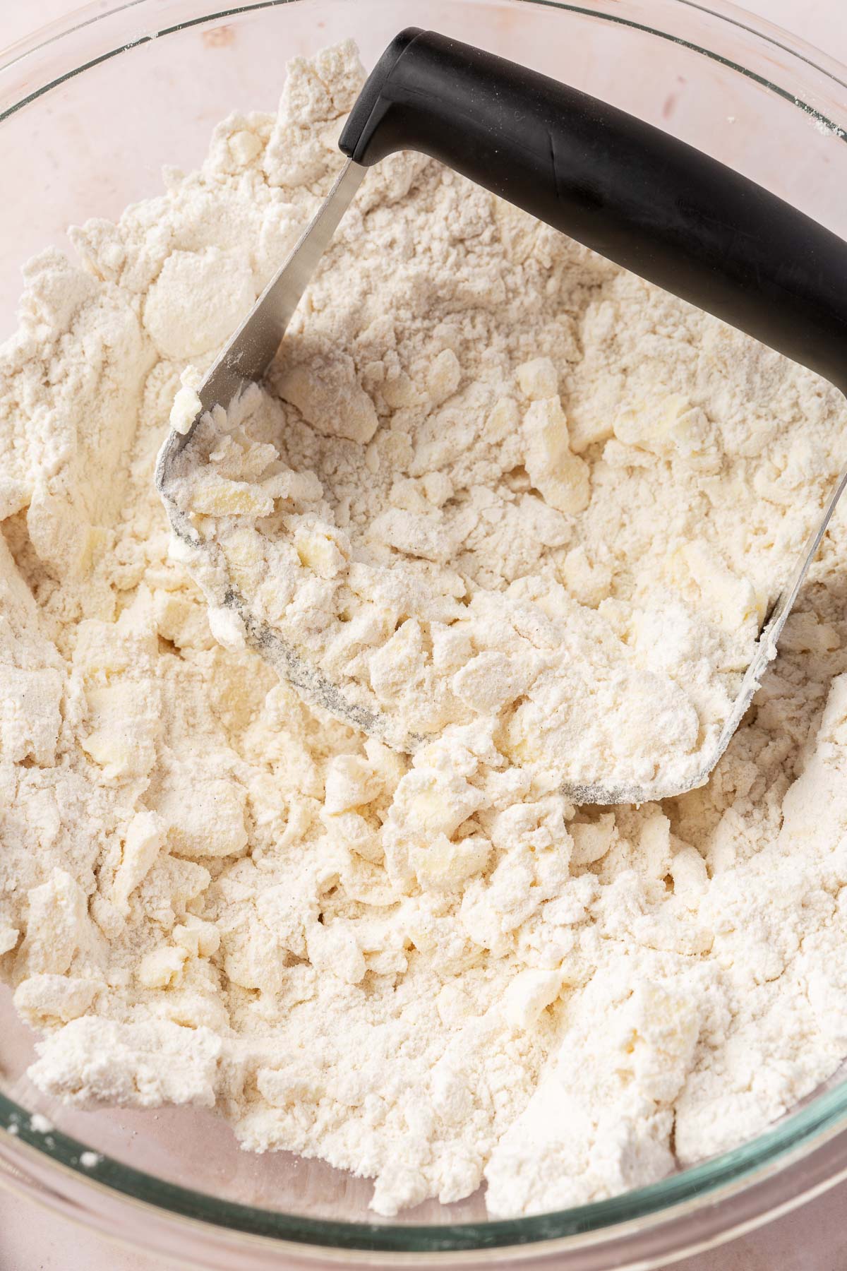 A pastry cutter that is cutting butter into a gluten-free flour mixture in a glass mixing bowl.