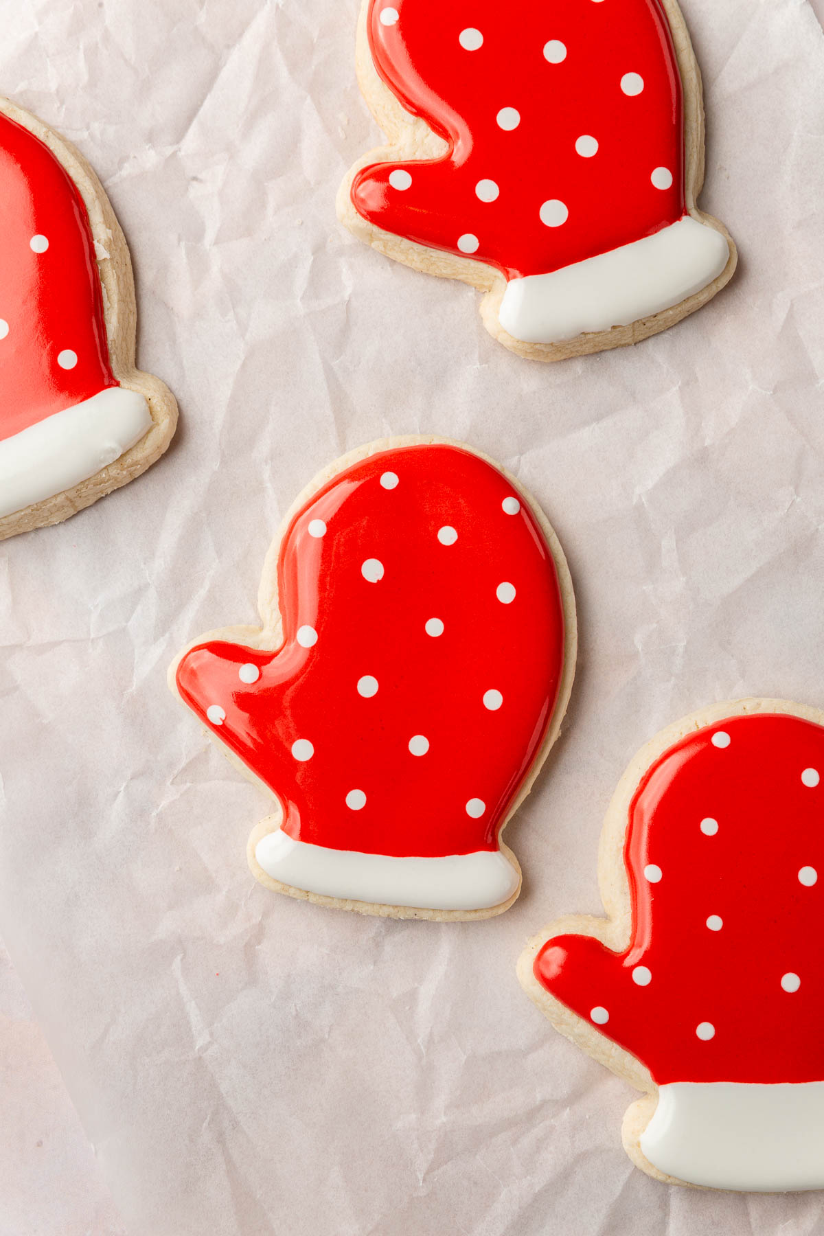 A few gluten-free mitten sugar cookies on a piece of parchment paper decorated with red and white royal icing with white polka dots using the wet-on-wet technique.