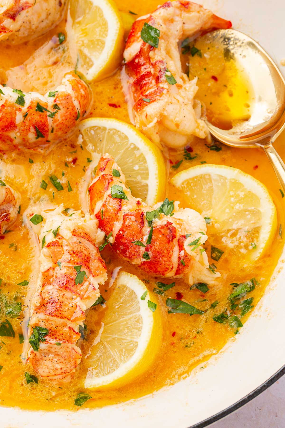 Six cooked lobster tails in butter sauce with garlic, parsley, and lemon wedges with a gold spoon for basting the lobster.