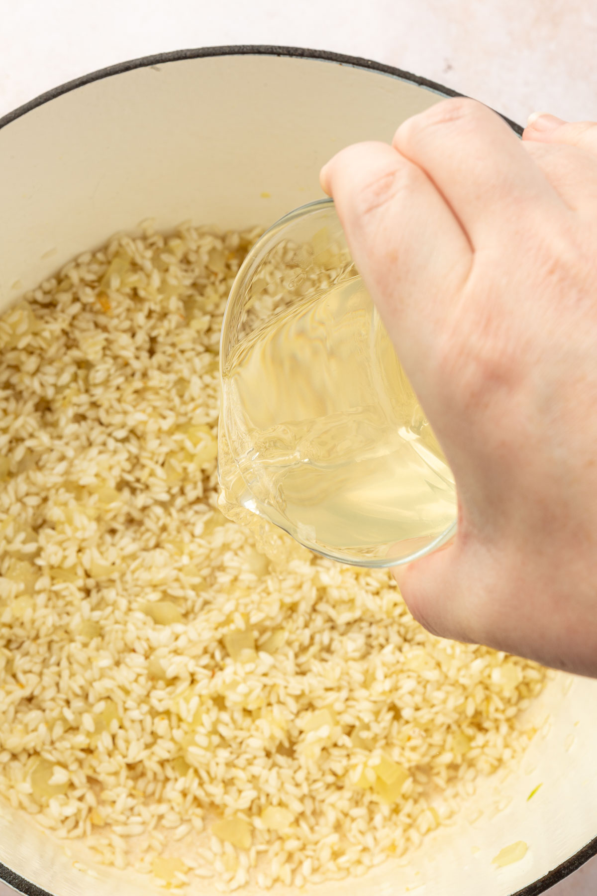 White wine being added to a white dutch oven of arborio rice and diced onions to make gluten free risotto.