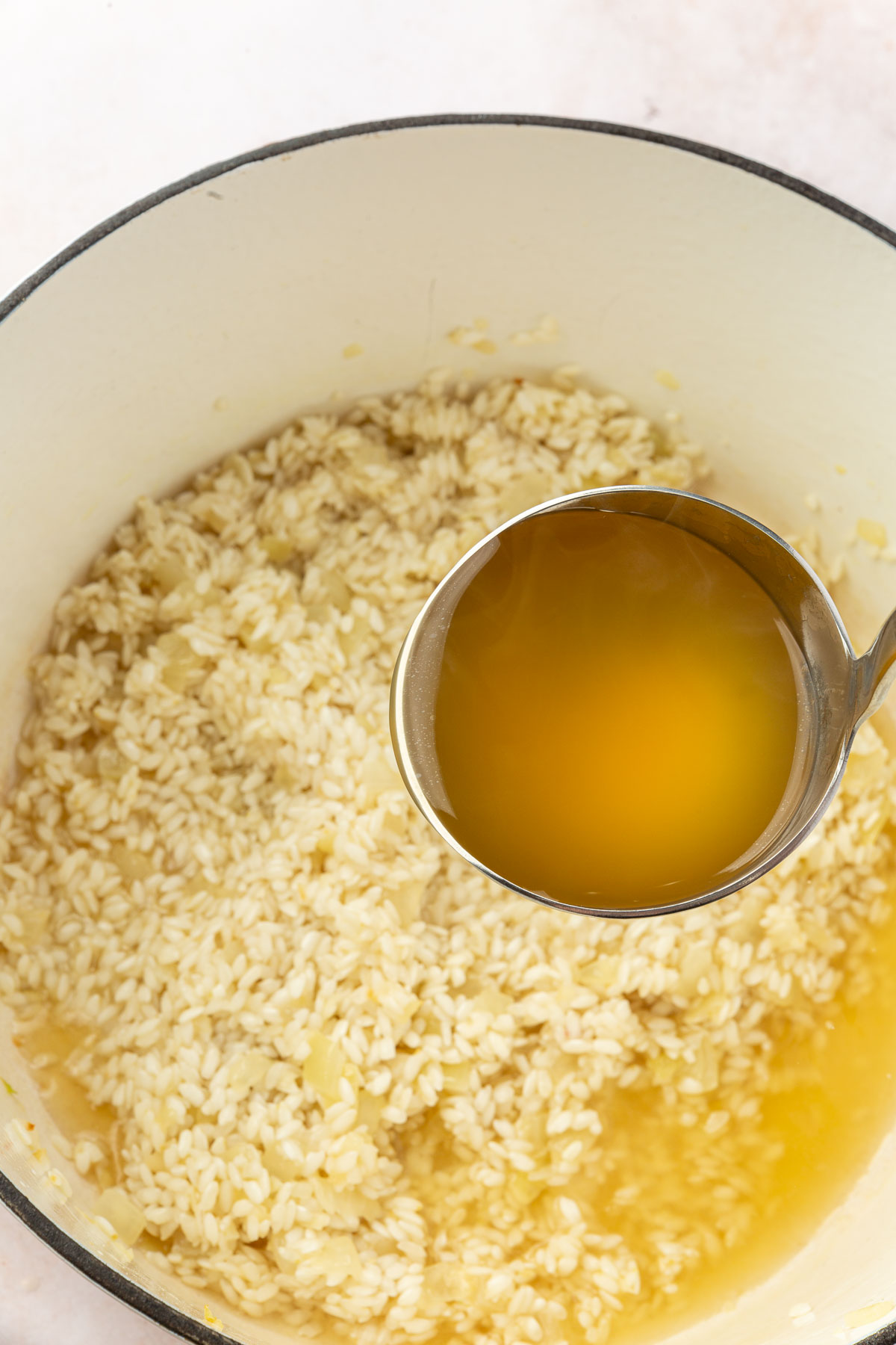 A ladle of chicken broth being added to arborio rice to make gluten free risotto in a white dutch oven.