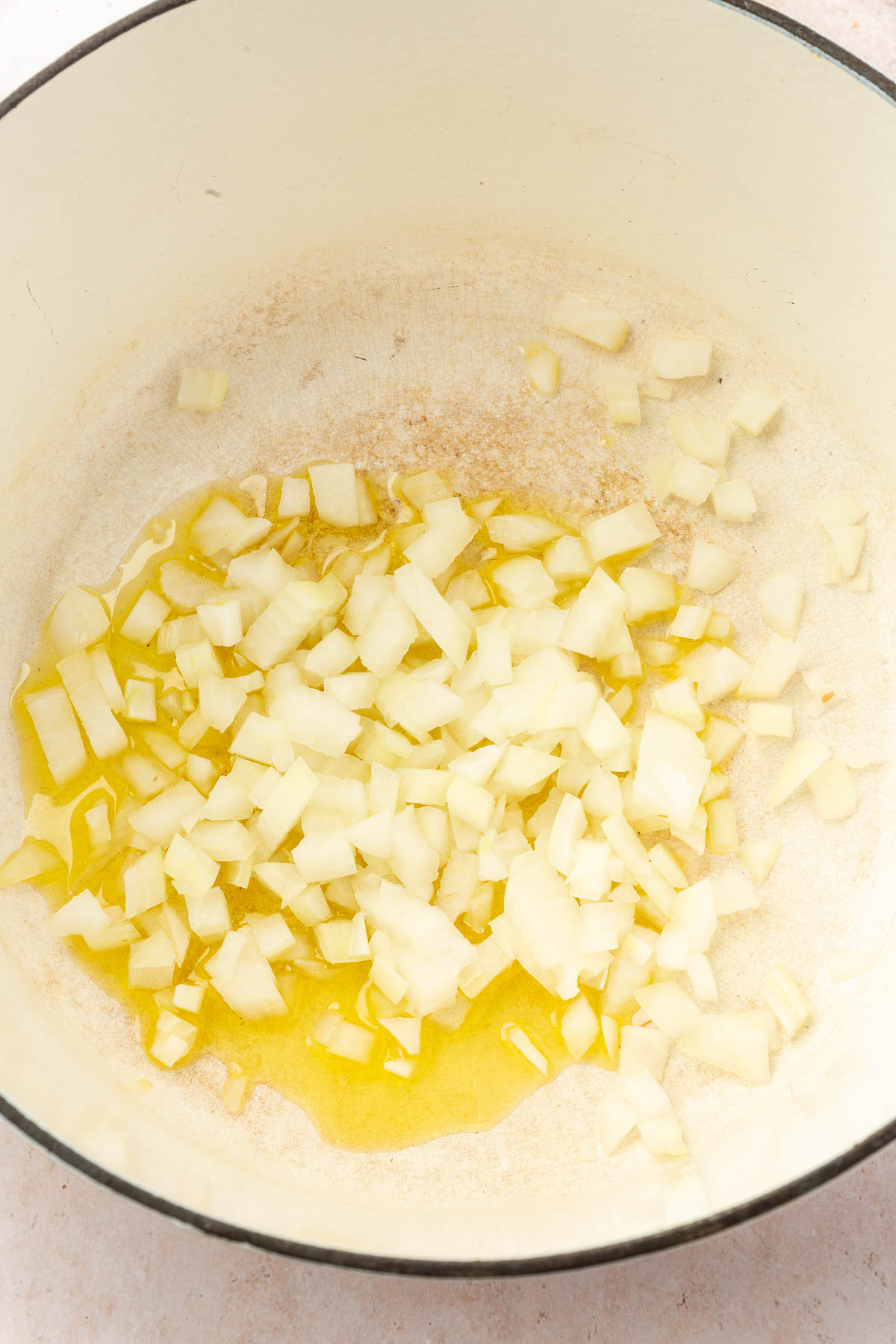 A white dutch oven with olive oil and raw diced onion before cooking.