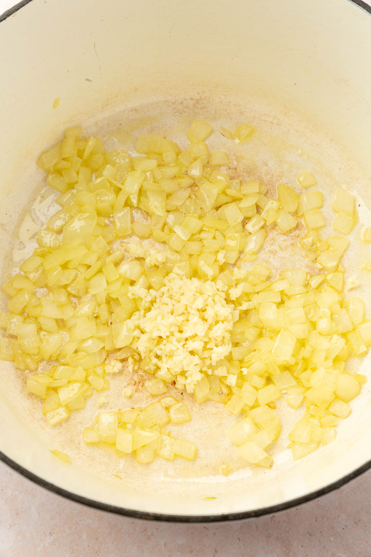 A white dutch oven with sautéed onion topped with minced garlic before mixing together.