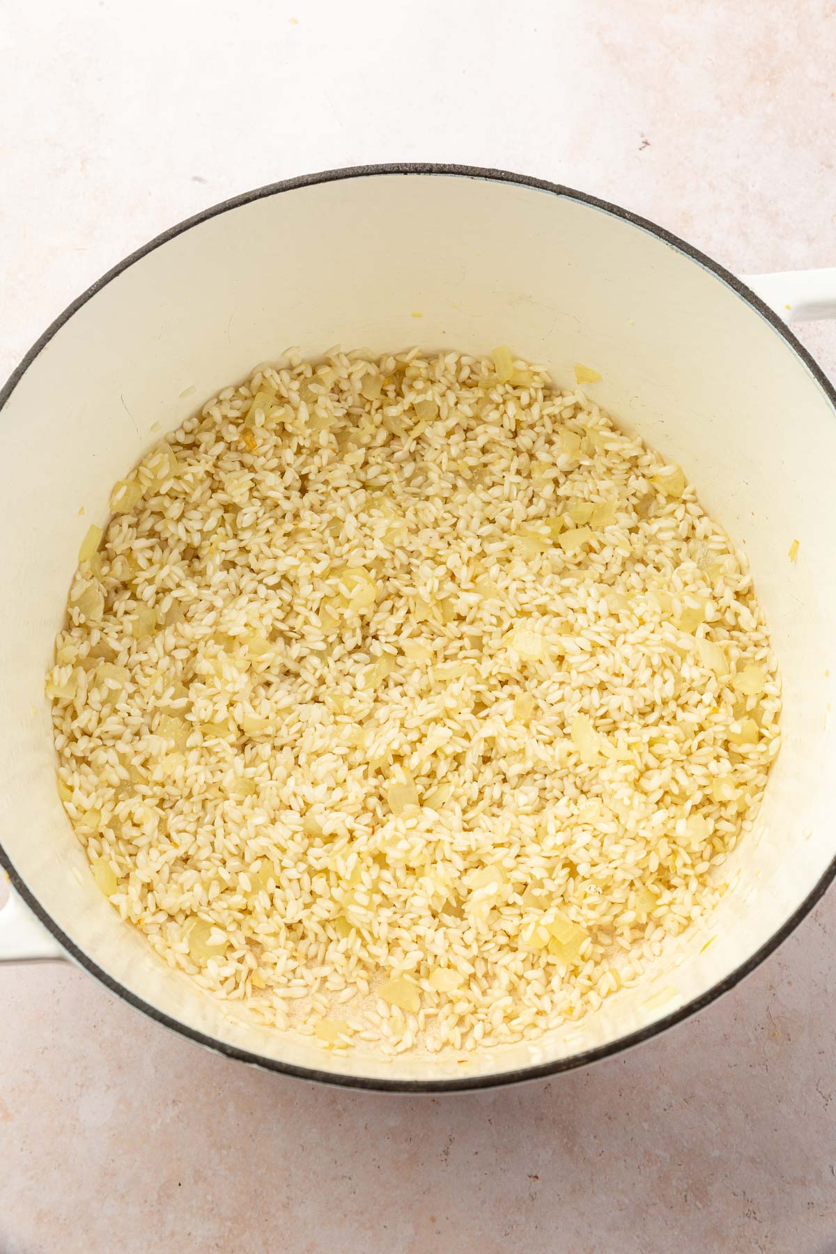 A white dutch oven with toasted arborio rice and sautéed onions to make gluten free risotto.