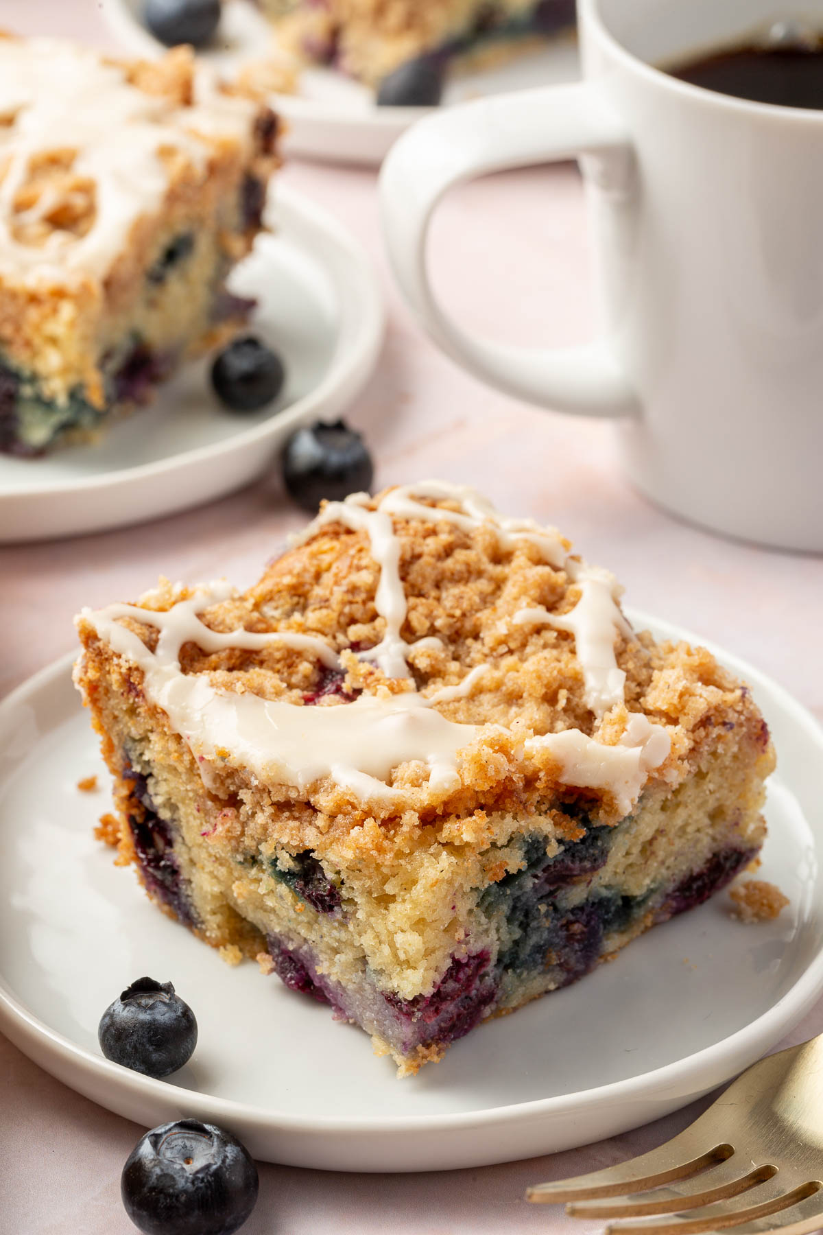 A slice of gluten-free blueberry coffee cake on a dessert plate with a mug of hot coffee in the background.