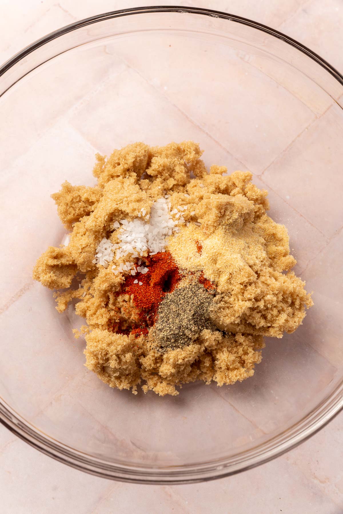 An overhead view of a glass mixing bowl with brown sugar, salt, garlic powder, pepper and paprika before mixing together.