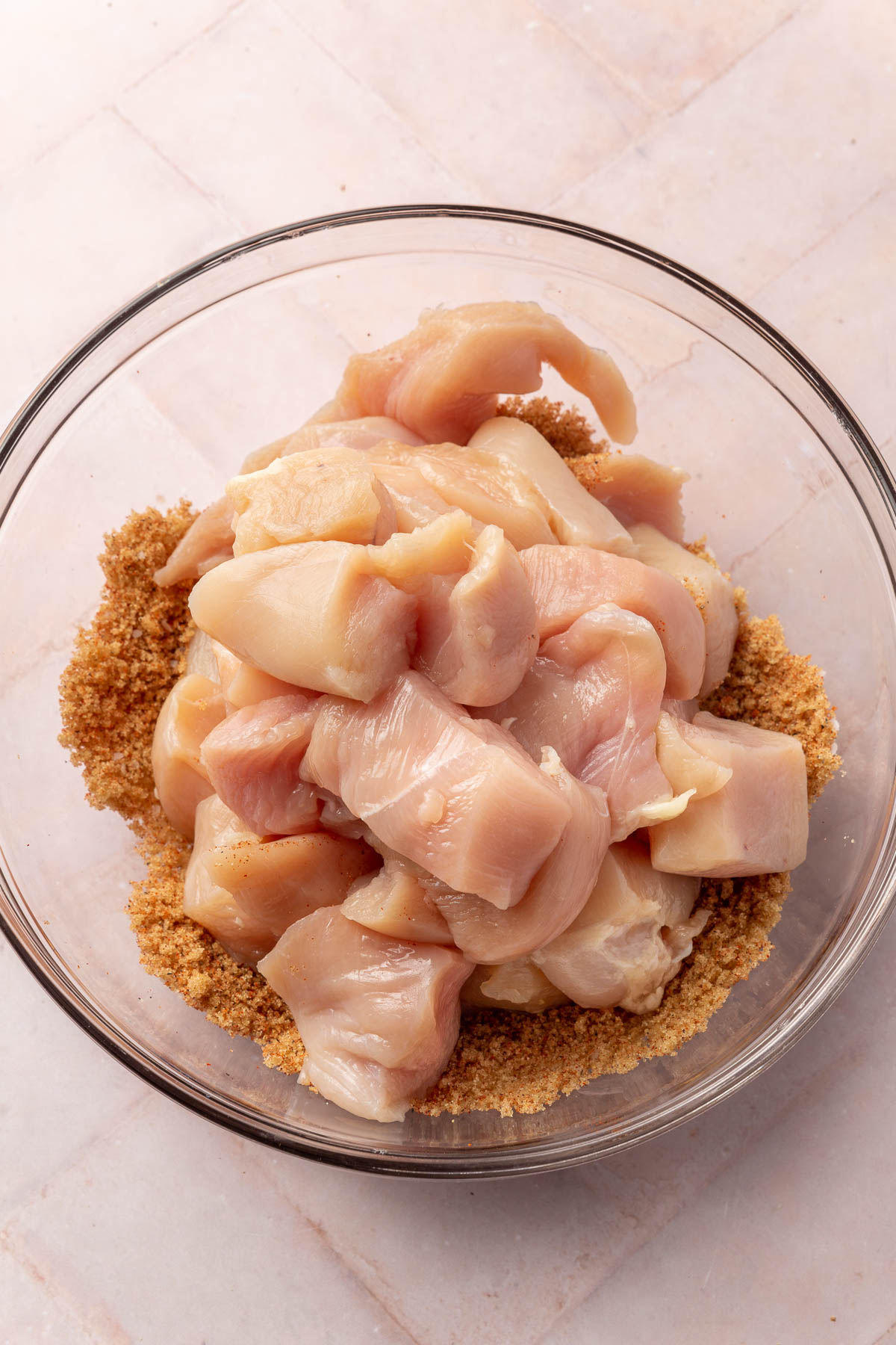 A glass mixing bowl with a pile of cubed chicken breast on top of a brown sugar mixture before mixing together.