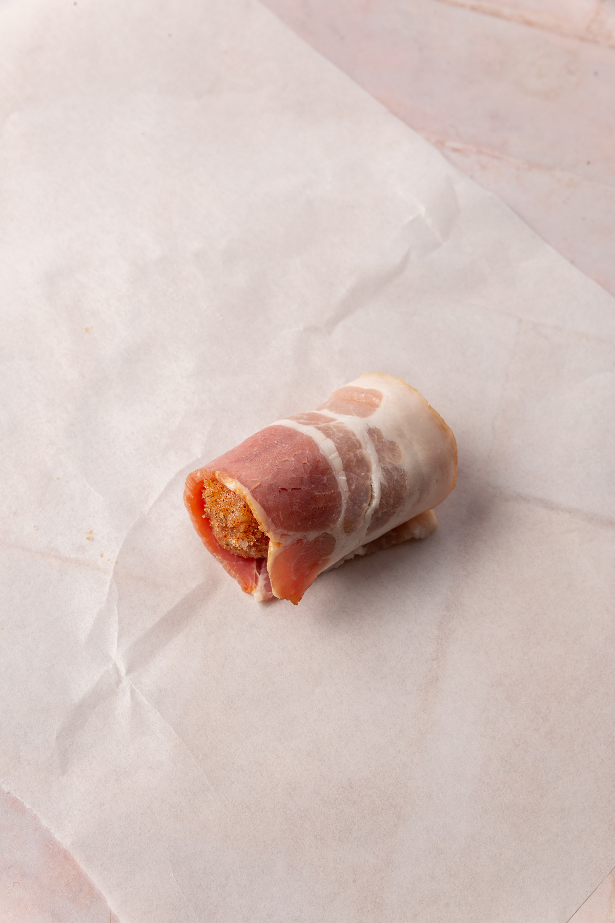 A single piece of brown sugar coated chicken rolled in bacon on a piece of parchment paper.