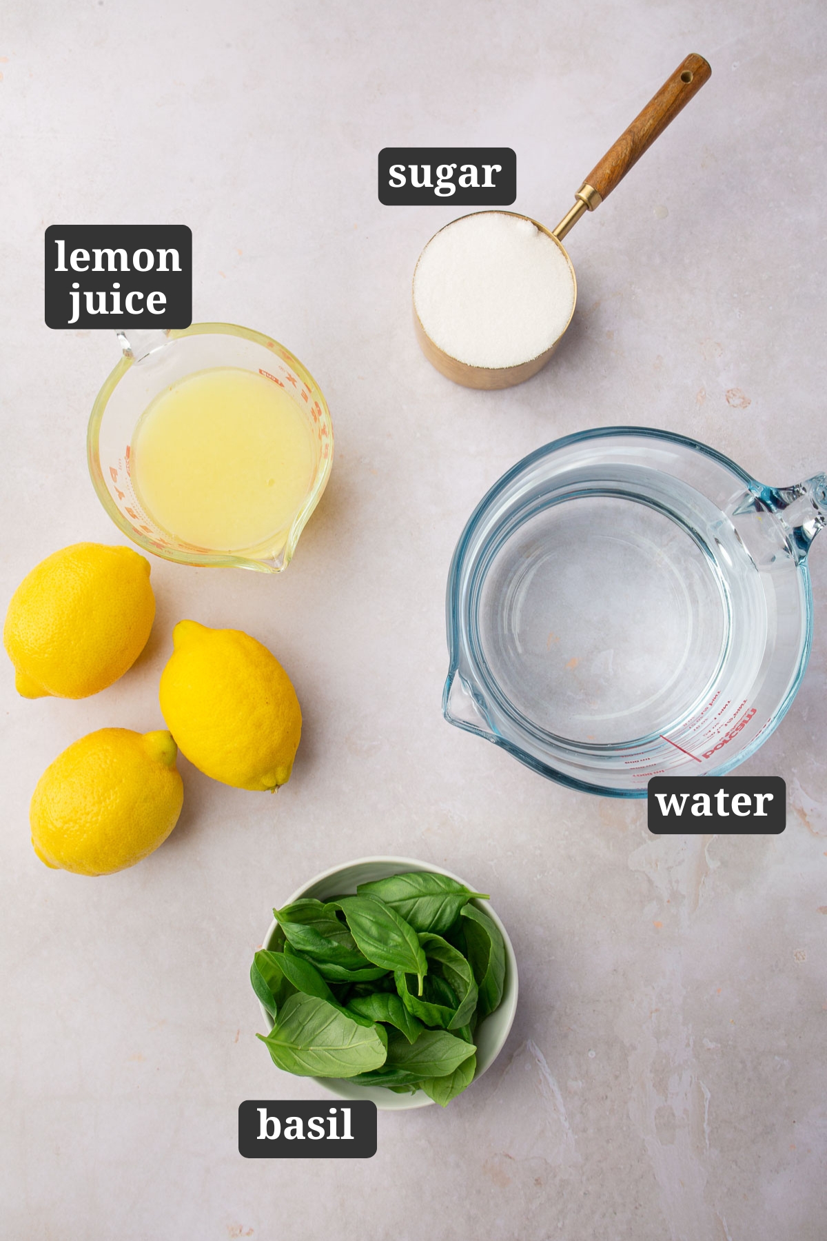 An overhead view of ingredients to make basil lemonade on a table including granulated sugar, fresh lemon juice, water, and fresh basil leaves with text overlays over each ingredient saying what it is.