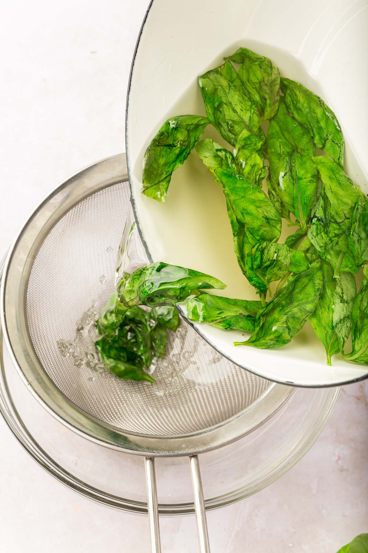 A saucepan pouring basil simple syrup into a wire mesh strainer to strain out the fresh basil.