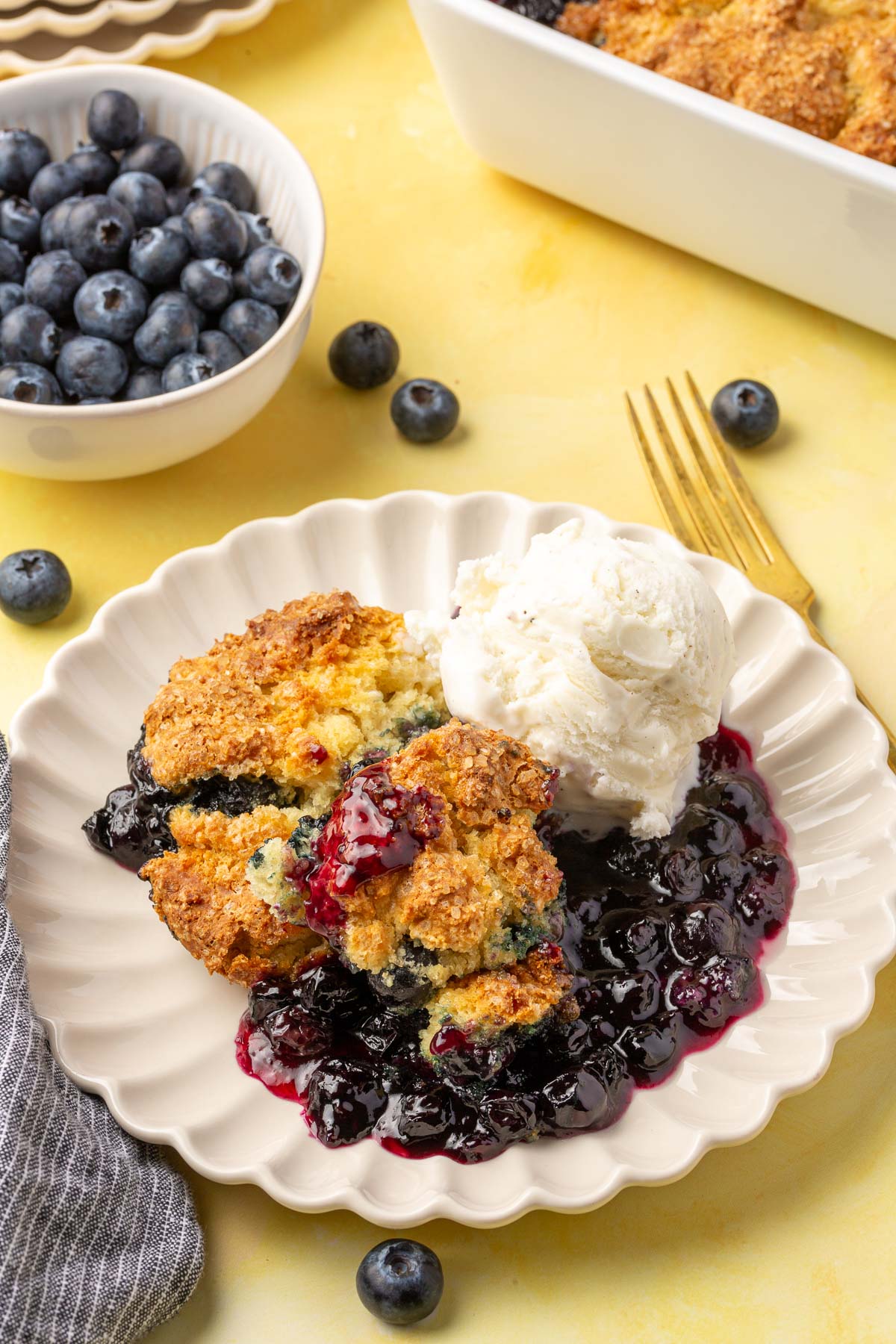 A single serving of gluten free blueberry cobbler on a scalloped plate with a scoop of vanilla ice cream with a bowl of fresh blueberries off to the side.