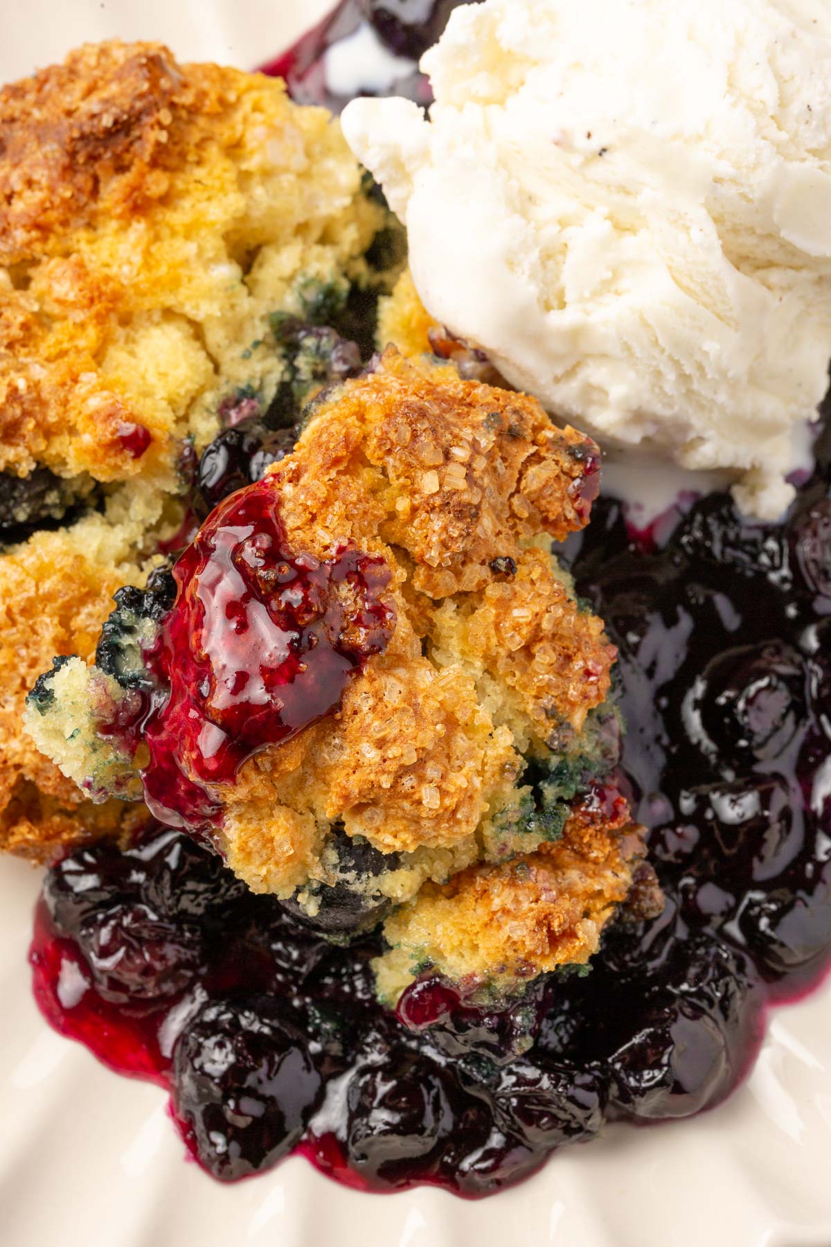 A close up of blueberry cobbler filling topped with a golden gluten free biscuit and a scoop of vanilla ice cream.