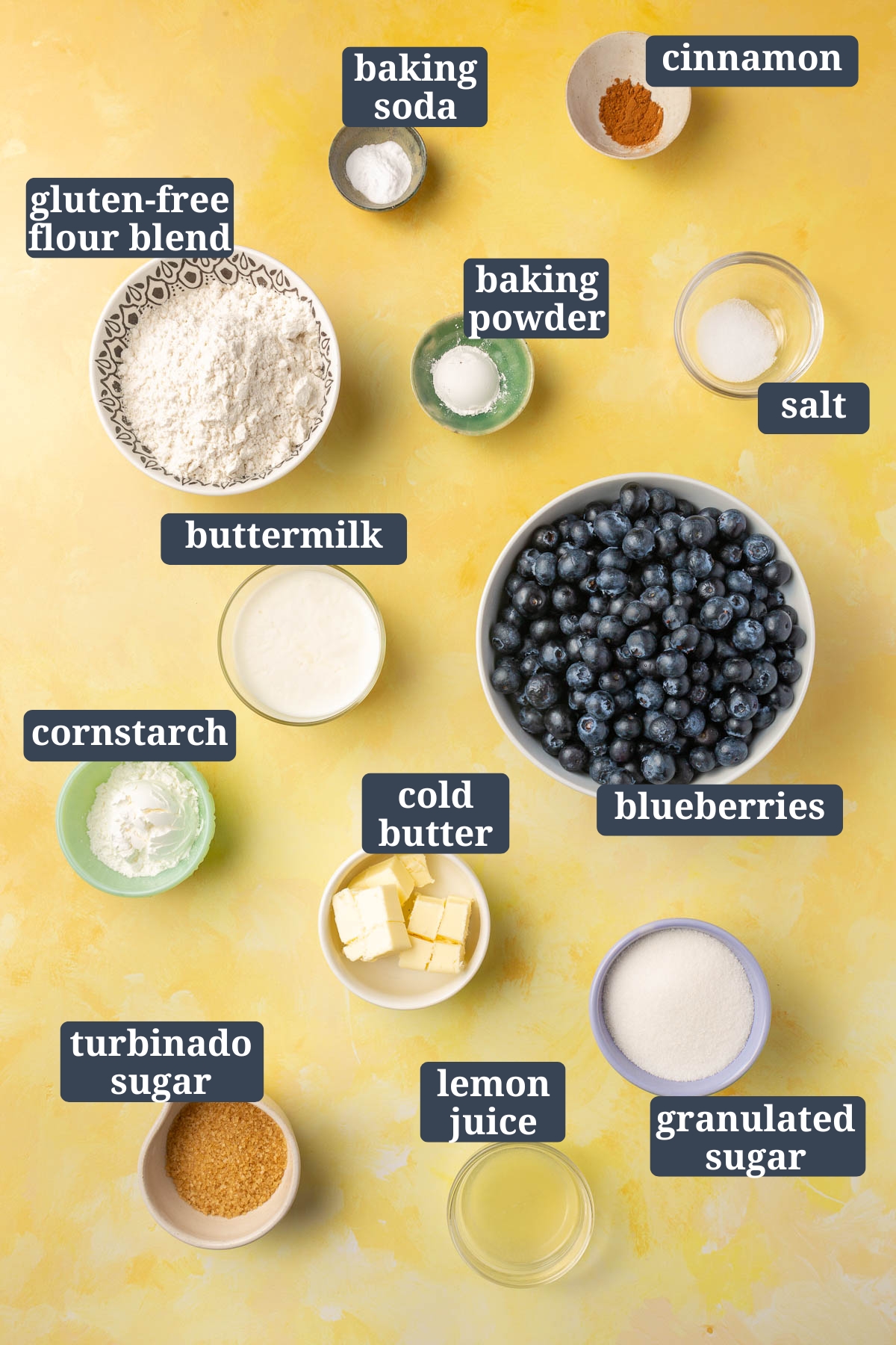Ingredients in small bowls to make gluten-free blueberry cobbler, including baking soda, baking powder, cinnamon, salt, gluten-free flour, buttermilk, blueberries, cornstarch, butter, granulated sugar, turbinado sugar, and lemon juice with text overlays over each ingredient to say what it is.