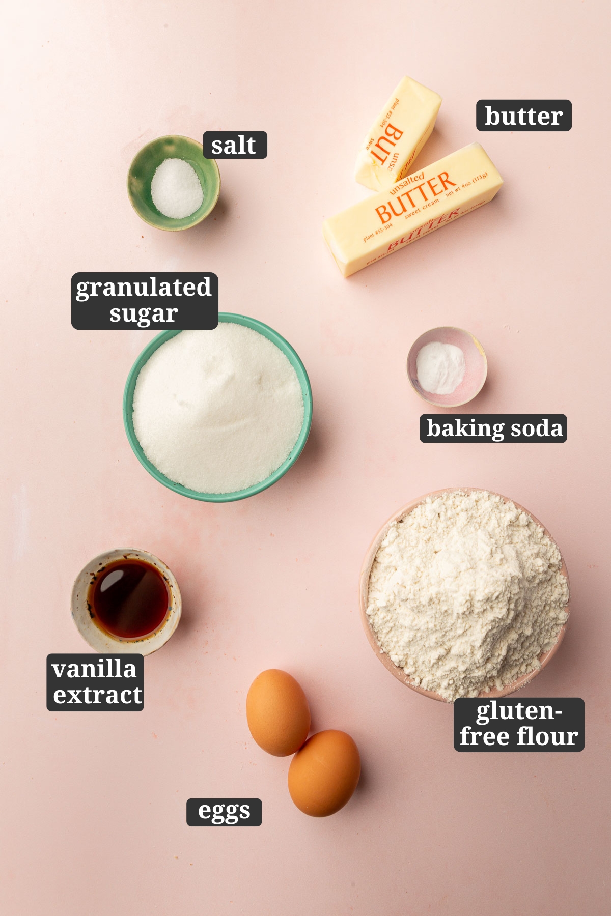 An overhead view of ingredients in small bowls to make gluten free chewy sugar cookies including unsalted butter, salt, baking soda, granulated sugar, vanilla, gluten-free flour and two eggs with text overlays over each ingredient.