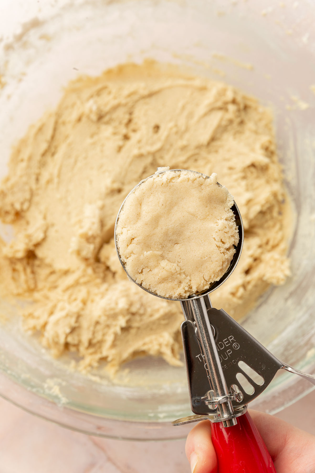A red portion scoop with a scoop of chewy sugar cookie dough in it over a bowl of additional gluten-free cookie dough.