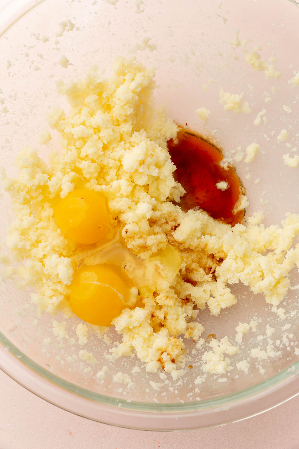 A glass mixing bowl with creamed butter and sugar topped with two raw eggs and vanilla extract before mixing together.