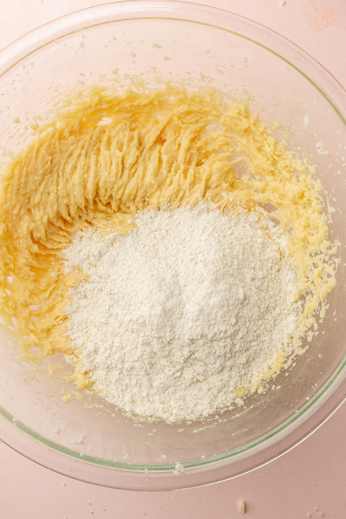A glass mixing bowl with creamed butter and sugar topped with gluten-free flour before mixing together.
