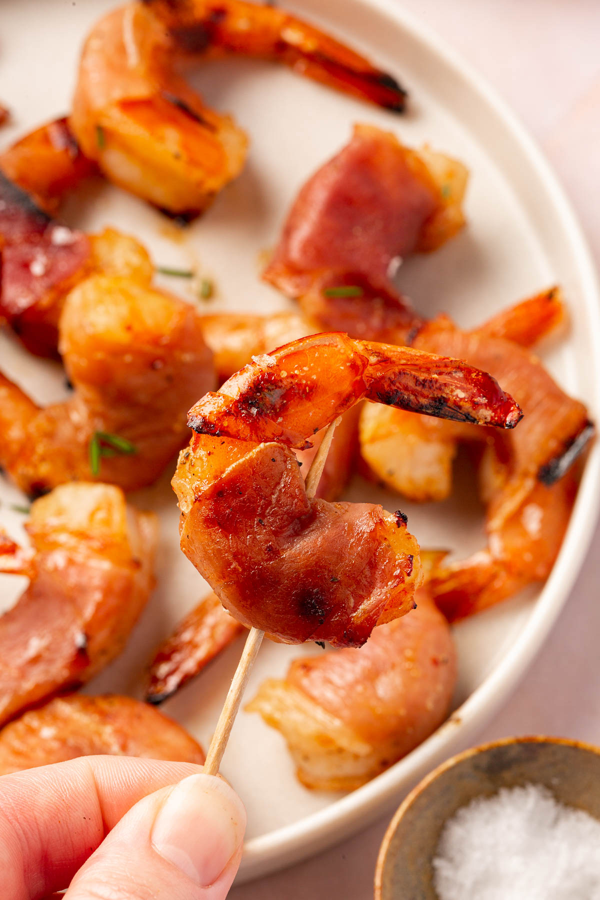 A hand holding a piece of prosciutto wrapped shrimp on a toothpick over a plate of additional shrimp.