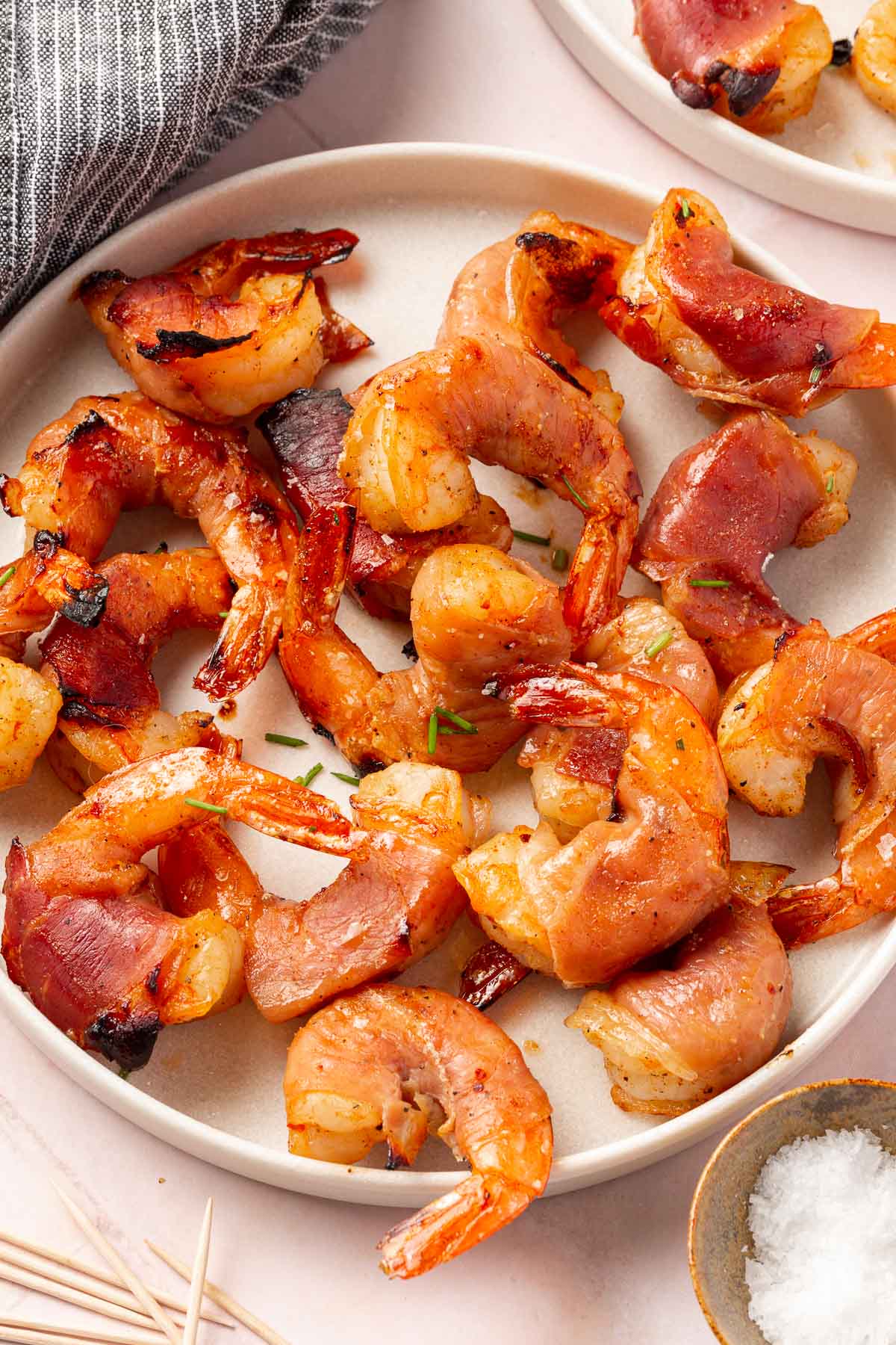 A serving plate of prosciutto wrapped shrimp that have been broiled in the oven with a bowl of flaky salt to the side.