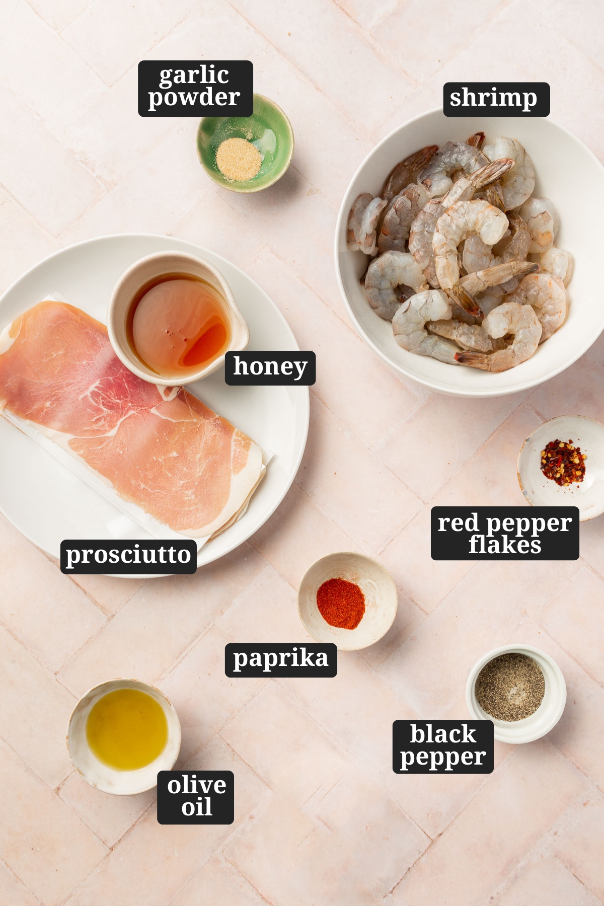 Ingredients in small bowls on a table to make prosciutto wrapped shrimp, including garlic powder, shrimp, honey, prosciutto, paprika, red pepper flakes, black pepper and olive oil with text overlays over each ingredient.