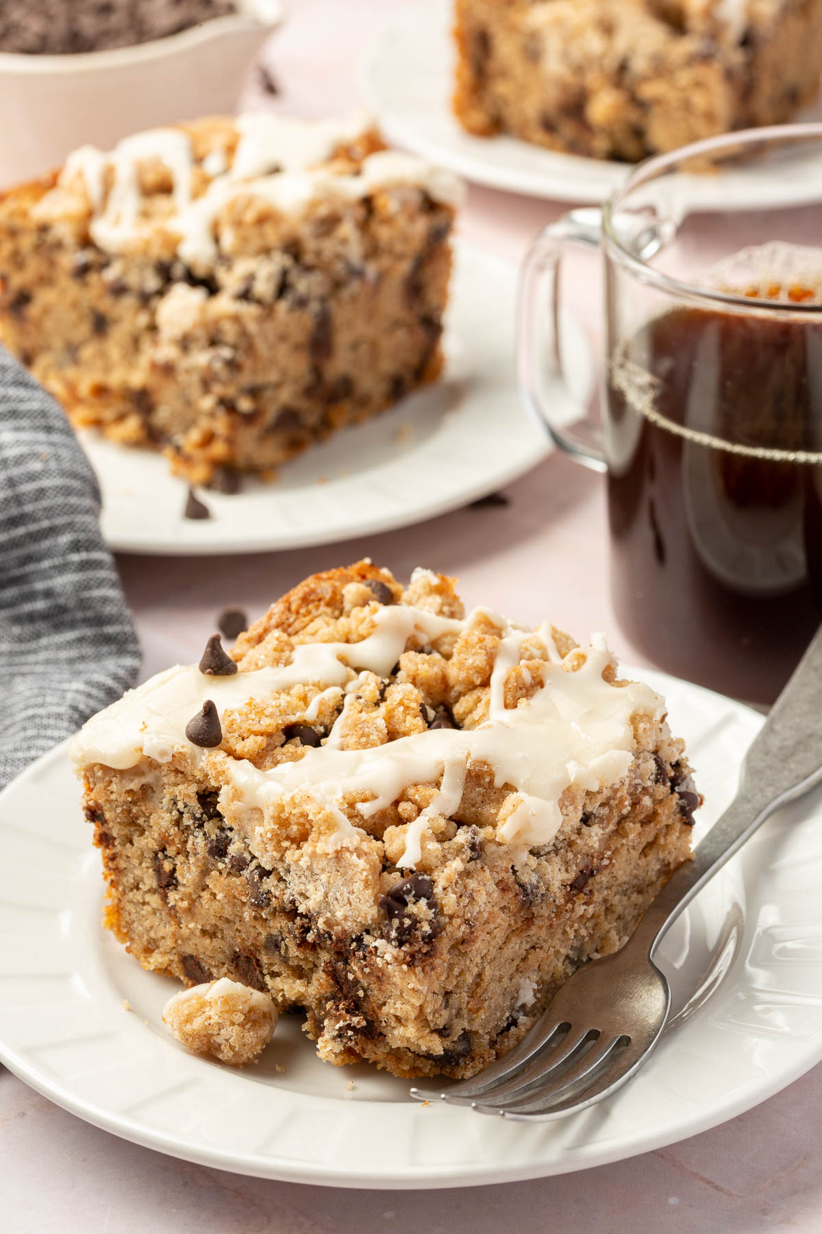 A slice of gluten free chocolate chip coffee cake on a dessert plate with a cup of coffee and additional slices of gluten free coffee cake in the background.