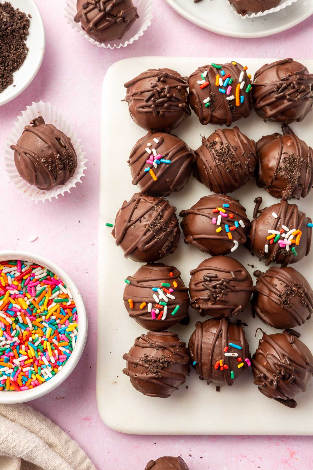 An overhead view of 15 Oreo truffles on a marble platter with additional truffles and rainbow sprinkles surrounding the platter.