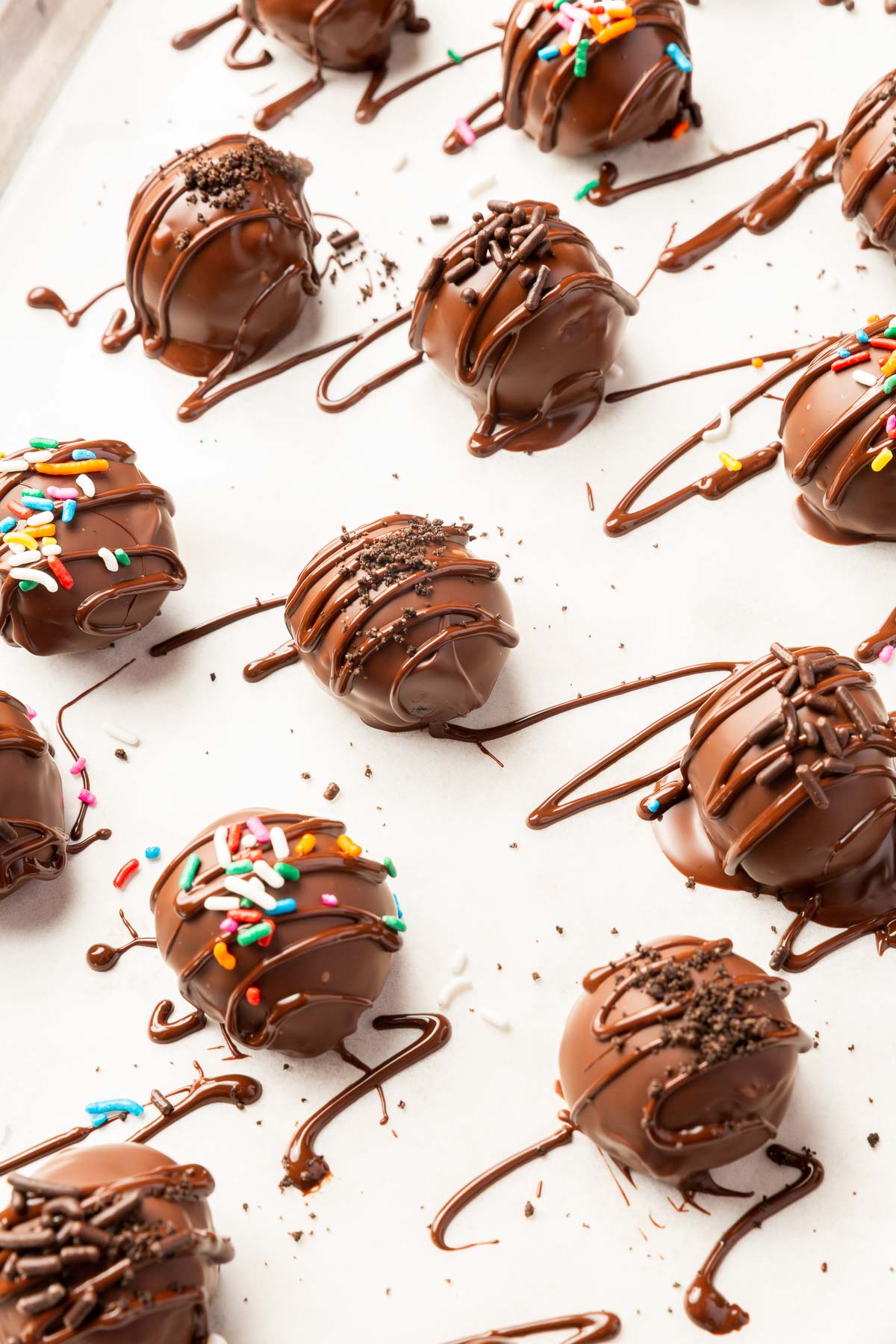 Gluten-free Oreo truffles dipped in melted chocolate and decorated with sprinkles on a piece of parchment paper with chocolate drizzles all over it.