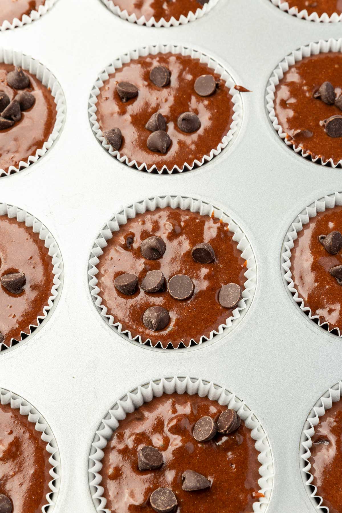 A muffin tin filled with chocolate batter, ready to bake gluten-free chocolate muffins, each topped with chocolate chips.