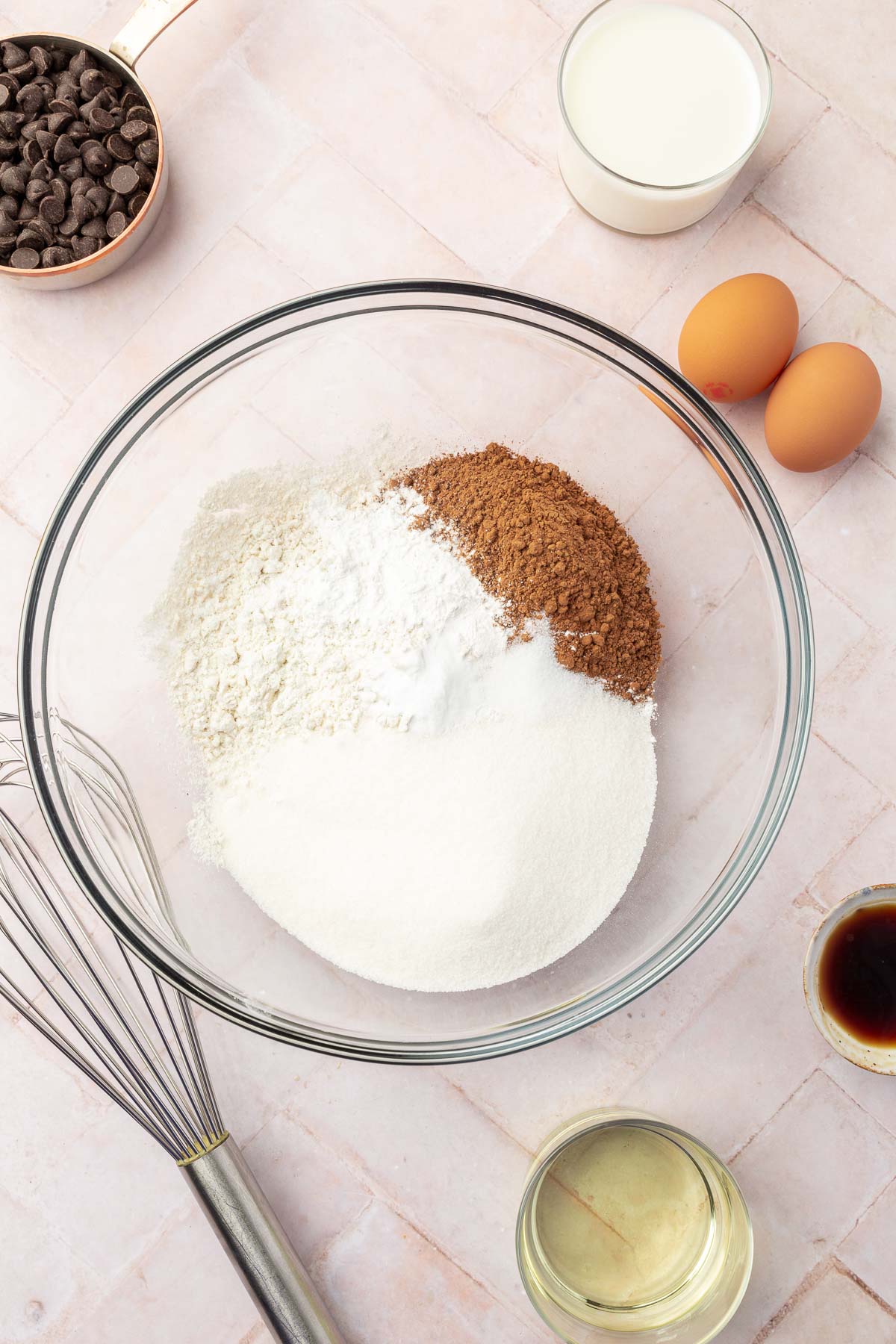 A mixing bowl with dry ingredients for gluten-free chocolate muffins, including gluten-free flour, sugar, cocoa powder, and baking soda, surrounded by other ingredients like eggs, milk, chocolate chips, and vanilla extract.
