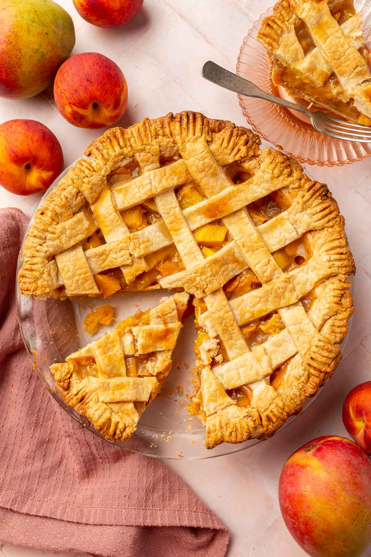 An overhead view of a gluten-free peach mango pie tat has been partially sliced with a single slice off to the side on a dessert plate and additional peaches and mangoes on the table.