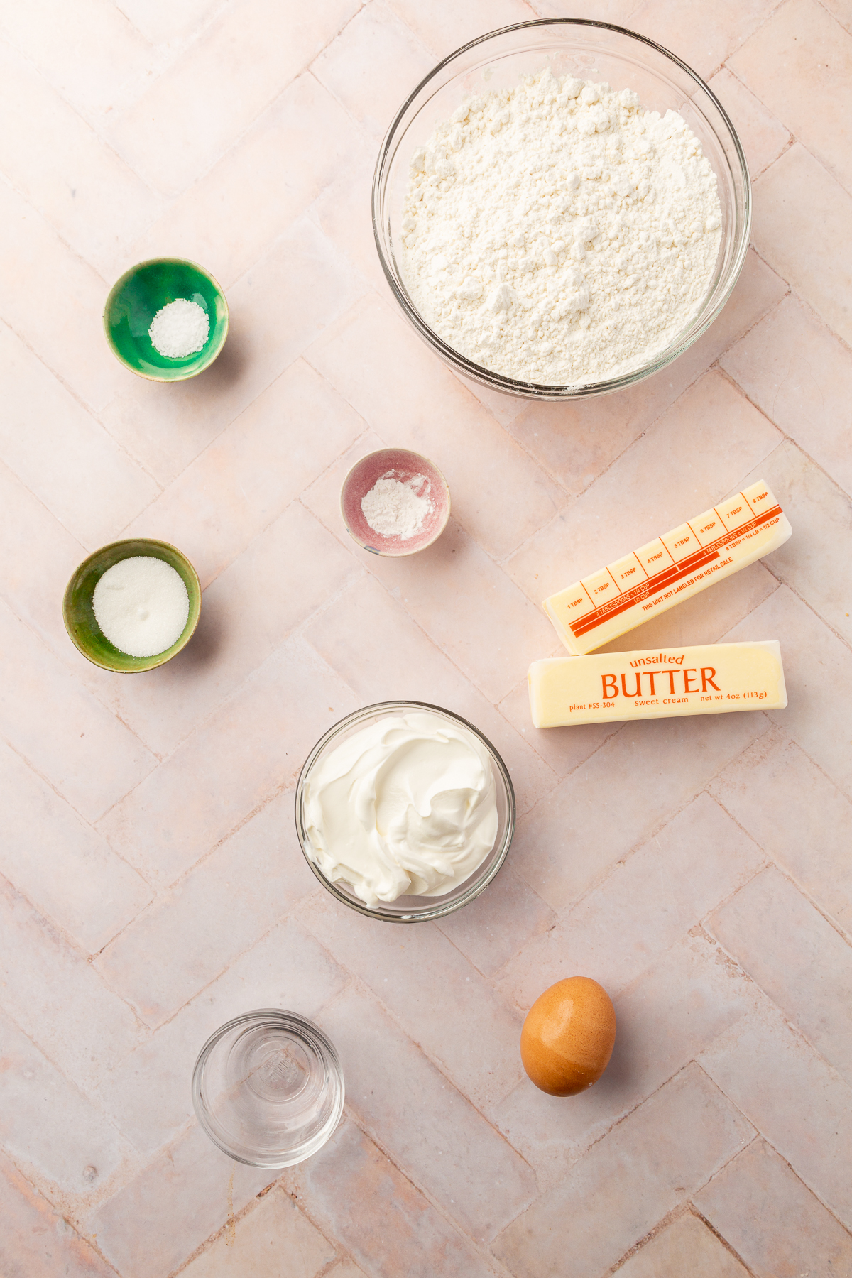 Overhead view of ingredients for gluten-free pie crust, including gluten-free flour, unsalted butter, sour cream, an egg, salt, sugar, and baking powder, arranged on a light-colored surface.