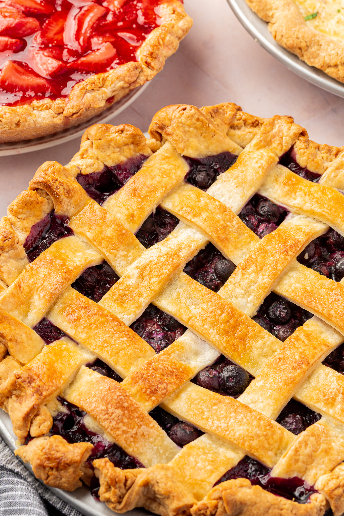 Close-up side angle of a gluten-free blueberry pie with a golden lattice crust, with another pie featuring a strawberry filling in the background.
