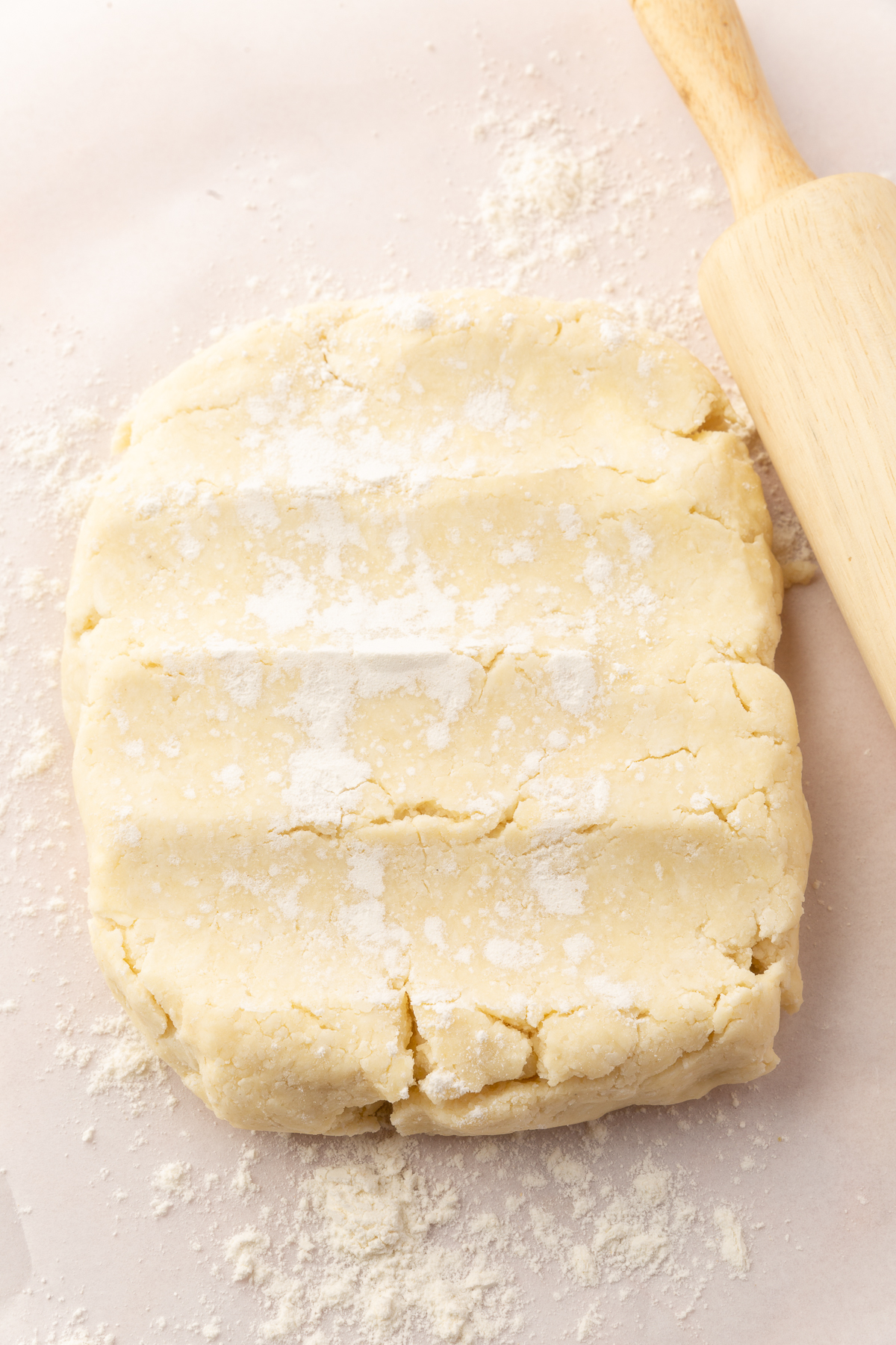 A flattened disk of gluten-free pie dough, lightly dusted with flour, rests on a sheet of parchment paper with a rolling pin nearby, ready to be rolled out.