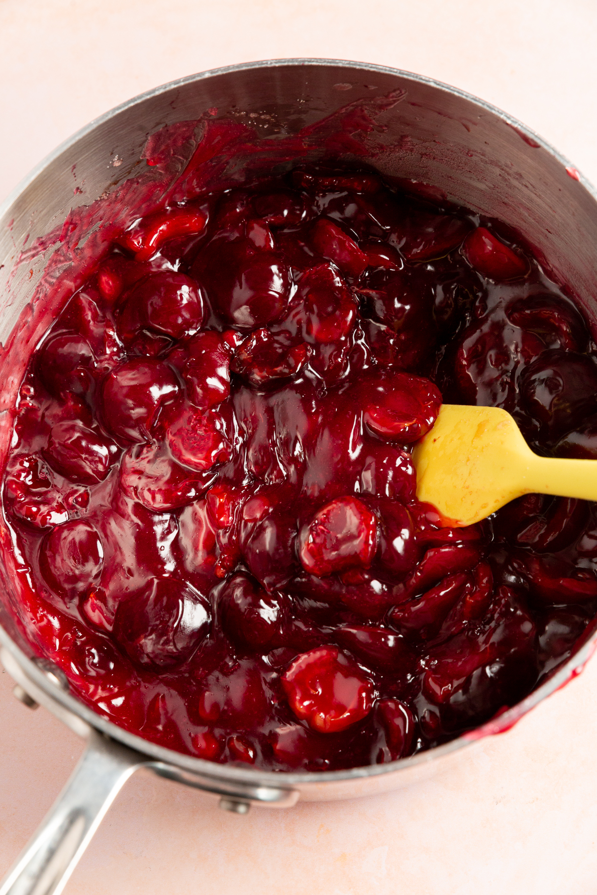 The cherry pie filling thickened and glossy, stirred with a yellow spatula in a saucepan.